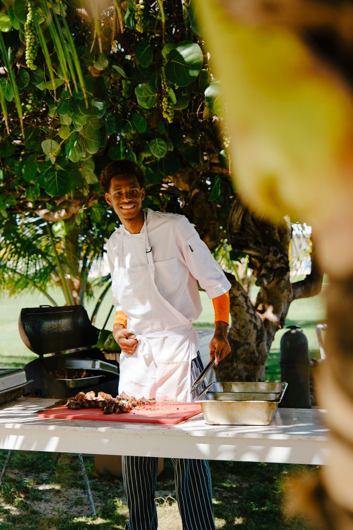 Person in chef's whites grilling food outdoors, smiling. Bright sunlight filters through trees; a table is set up.