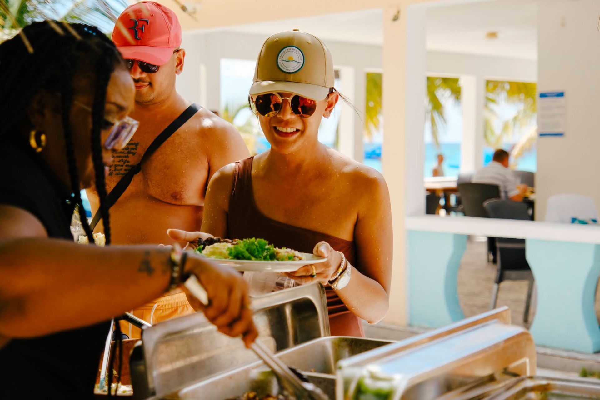 People serving food at an outdoor buffet. Bright setting, blue and white decor. Smiling woman receives food.