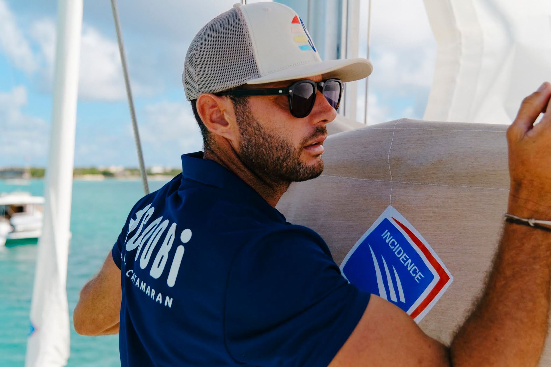 Man in sunglasses and hat on a sailboat, looking at something off-camera; wearing a blue shirt.