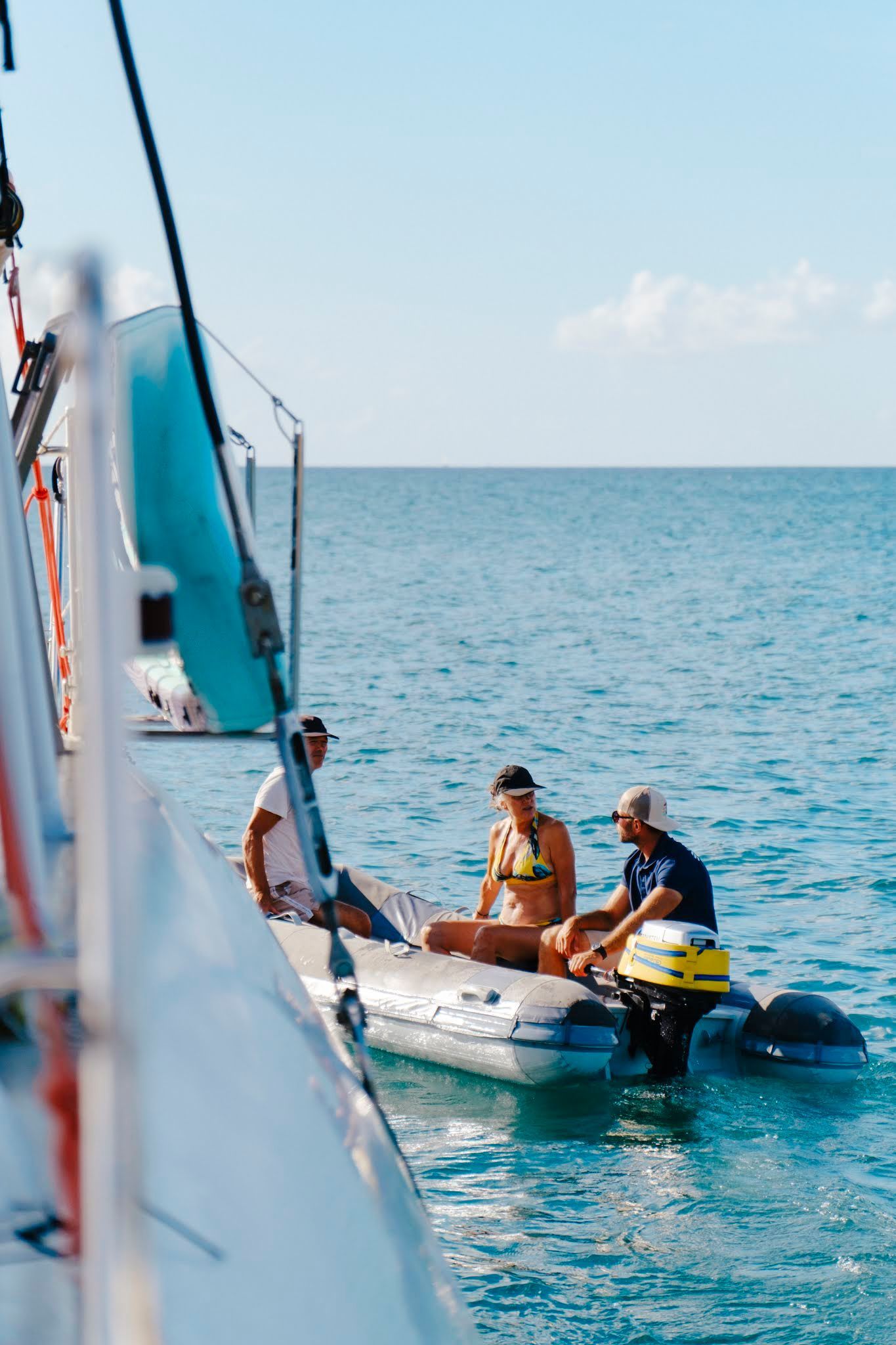 Three people in a small inflatable boat near a larger sailboat on a sunny day.
