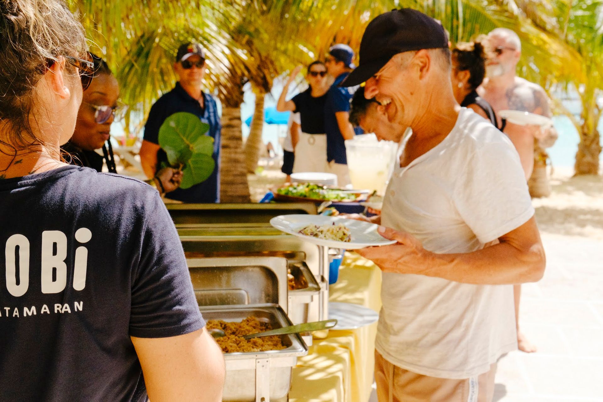 People at beach buffet; man with plate, smiles; food table, sunny setting.
