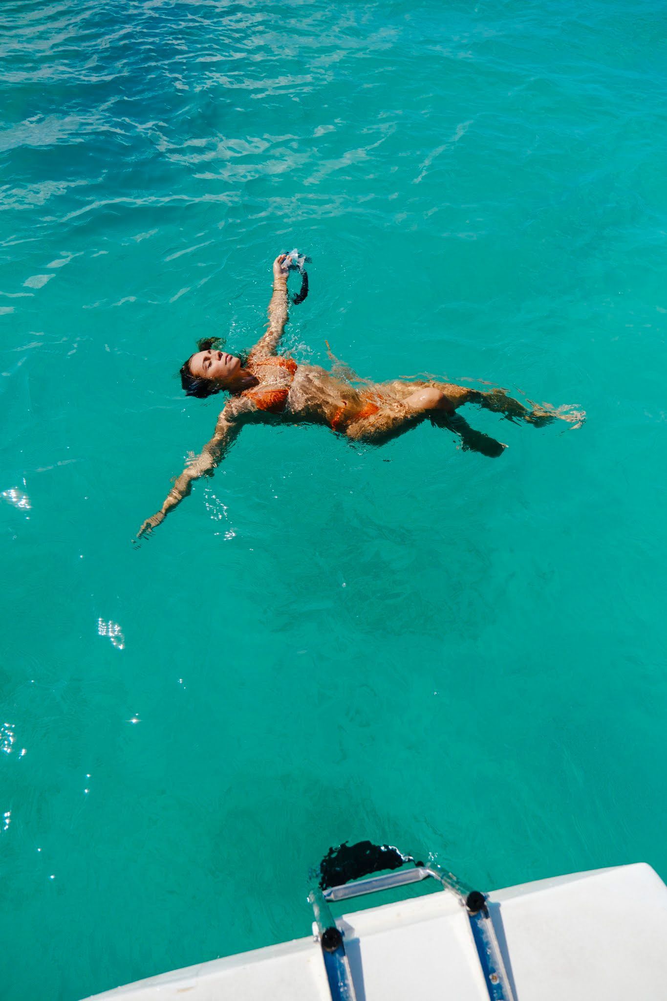 Person floating in turquoise water, wearing orange bikini, holding goggles. Boat deck in foreground.