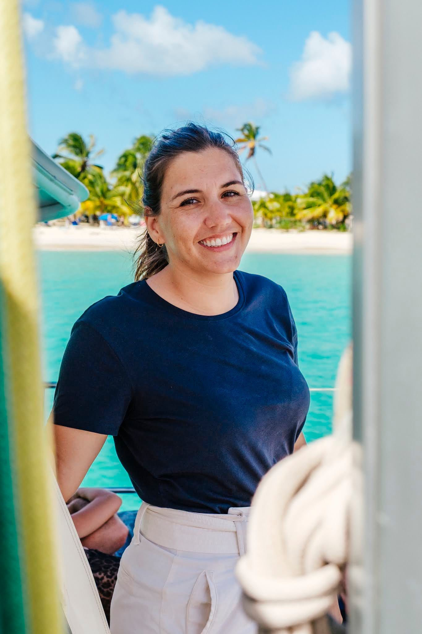 Woman on a boat smiles at the camera, with a tropical beach and ocean in the background.