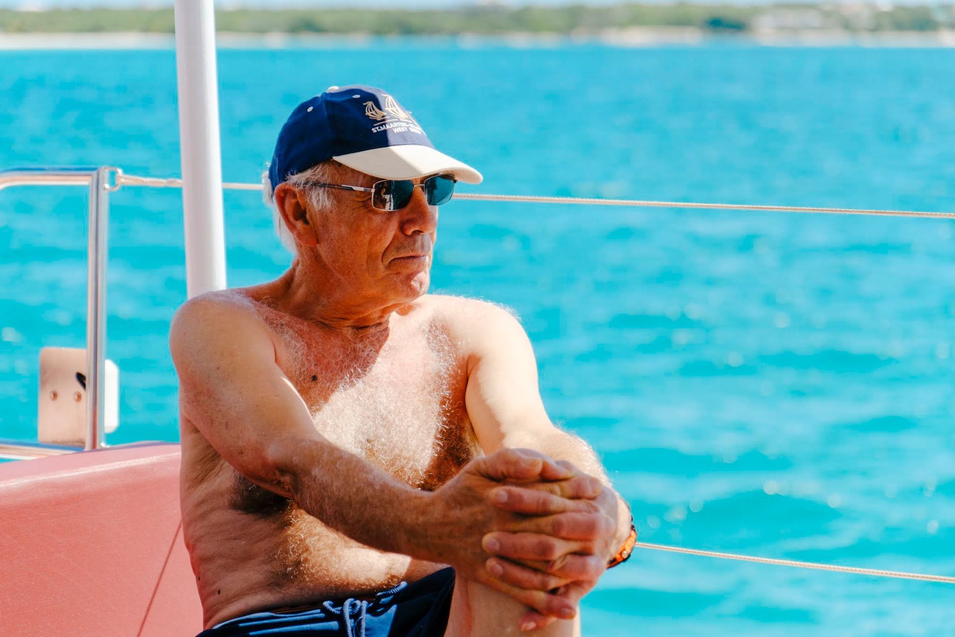 Un homme portant un chapeau et des lunettes de soleil, torse nu, est assis sur un bateau et contemple l'eau turquoise.