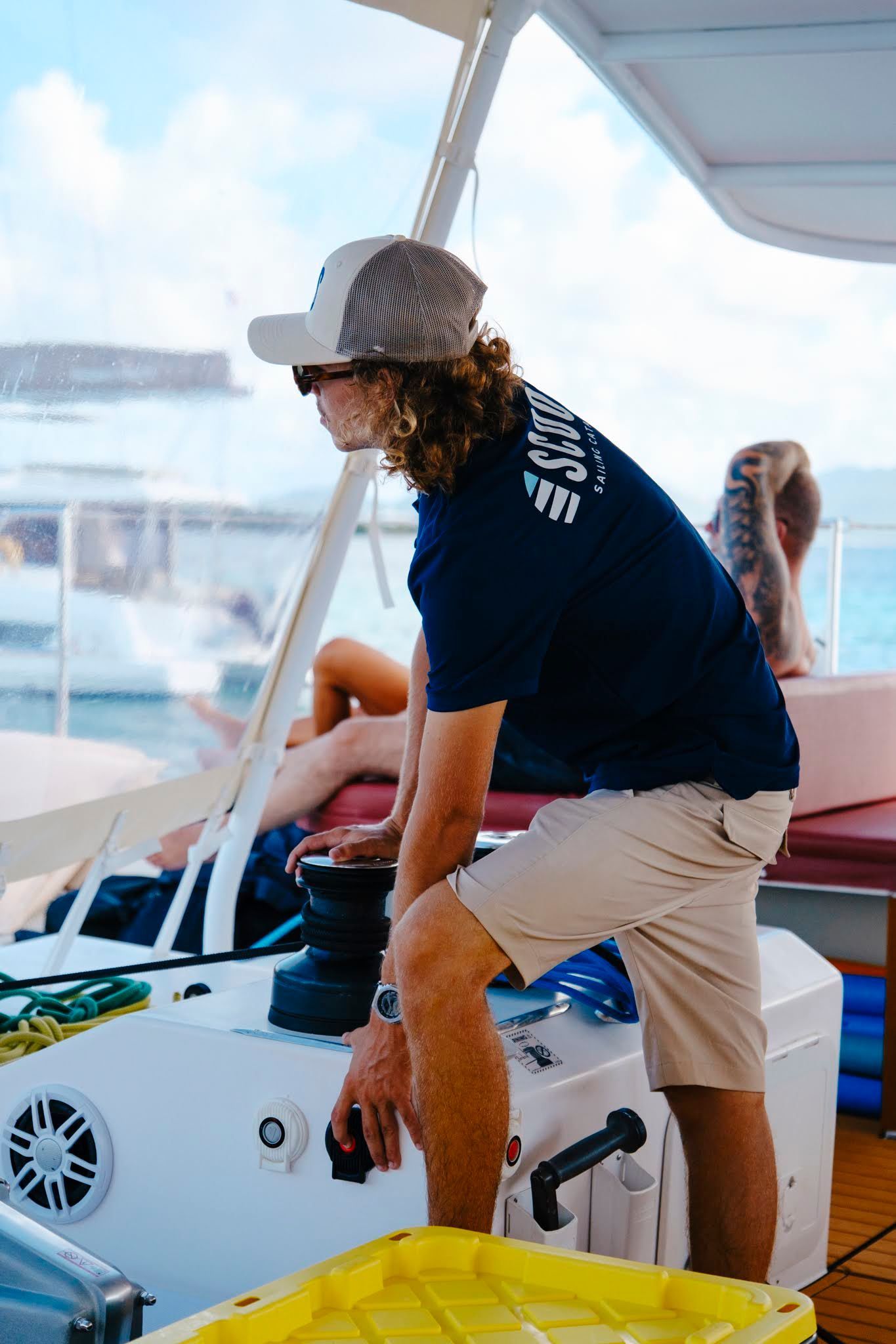 Man in hat and sunglasses operating boat controls; passengers relax in background.