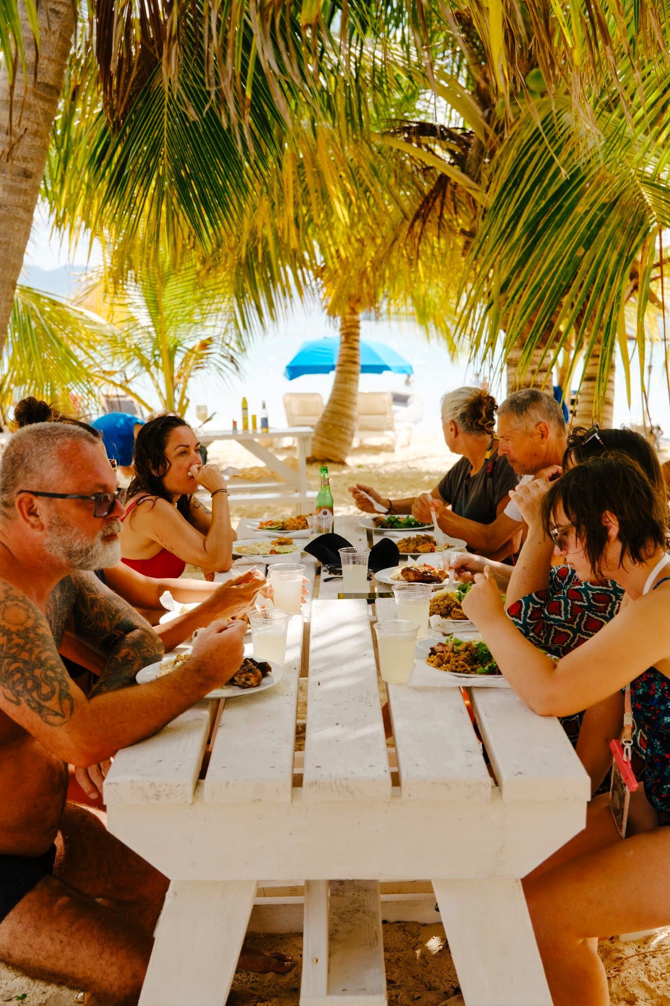 People dine at a long white table under palm trees on a beach.