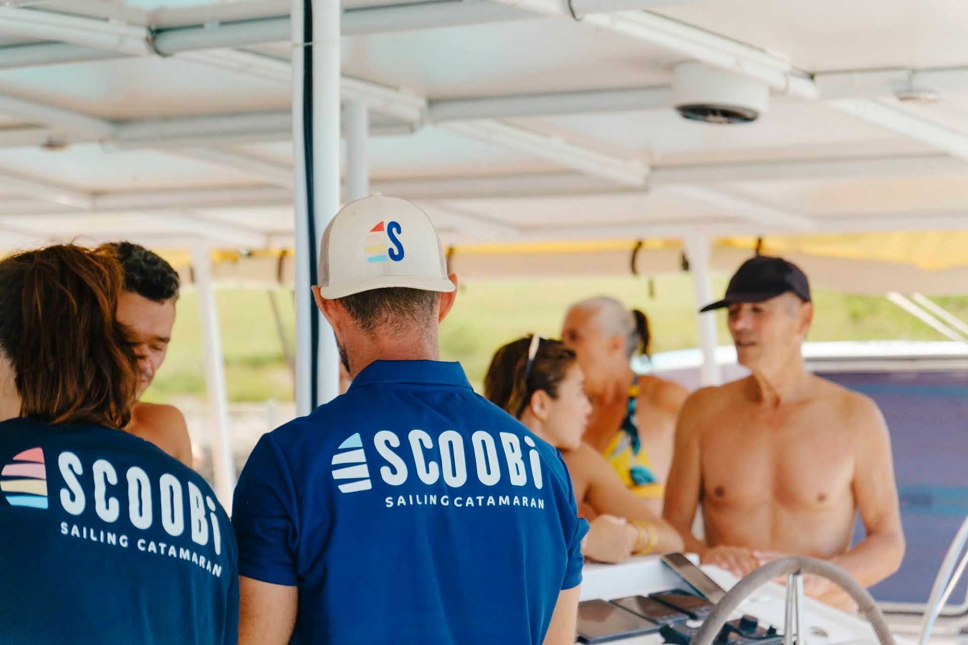 Des personnes sur un catamaran ; un homme porte une casquette et un t-shirt SCOOBI, d'autres regardent la mer.