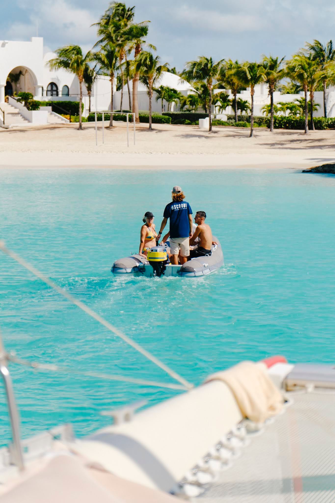 Small boat on turquoise water with three people and a motor, near a beach and white buildings.