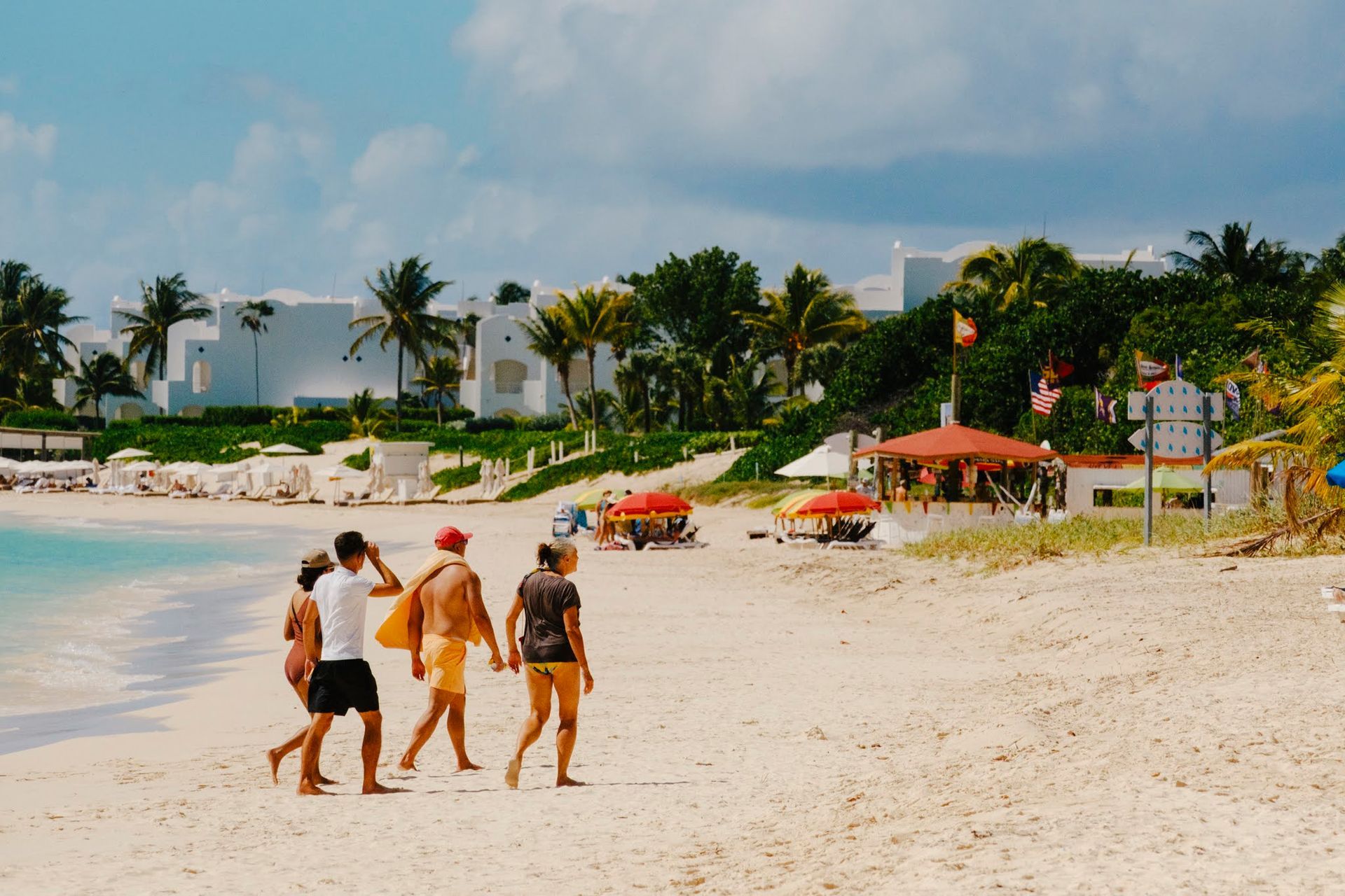 Des personnes se promènent sur une plage de sable fin ; eau turquoise, bâtiments blancs et parasols en arrière-plan sous un ciel bleu.