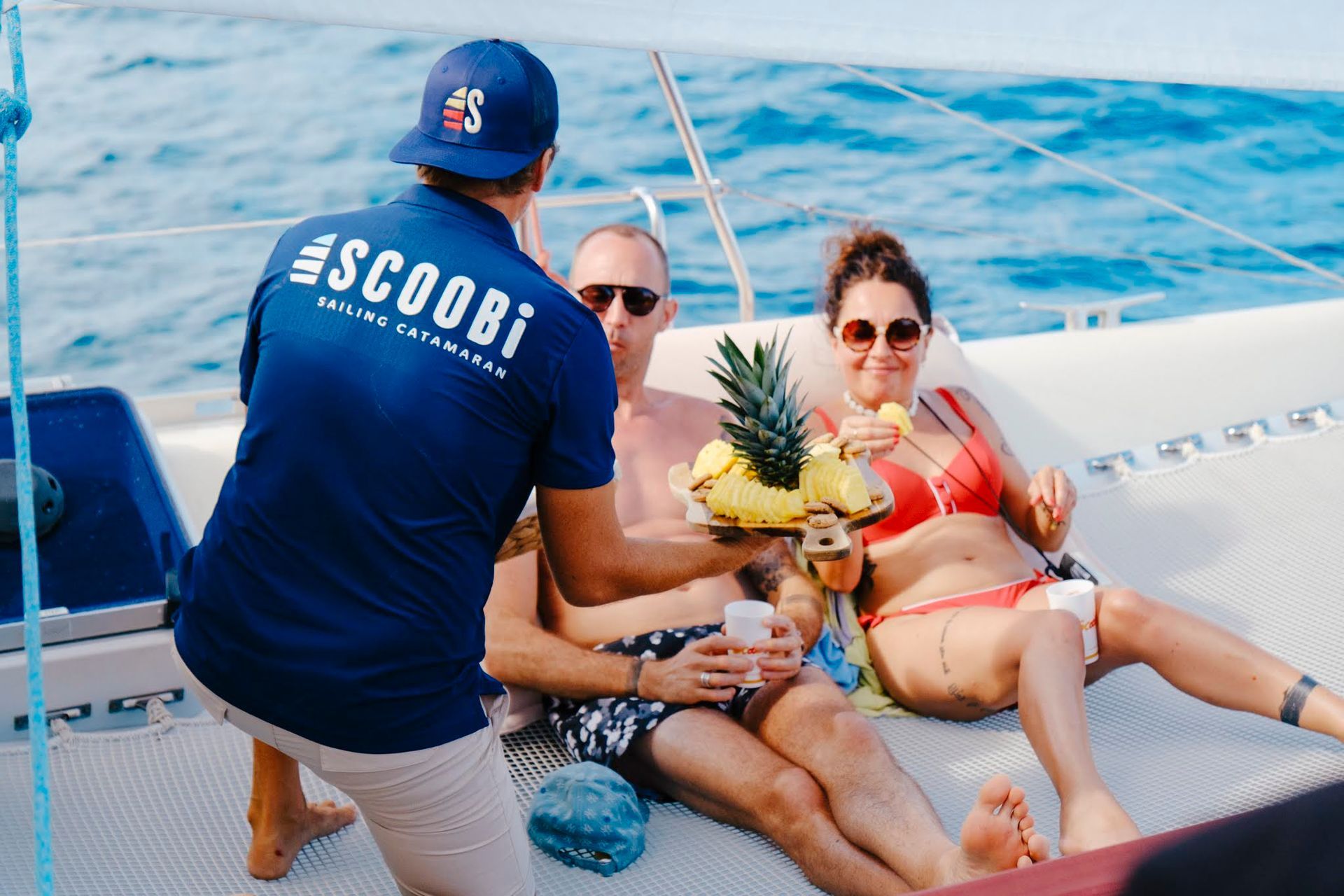 Waiter serving drinks and fruit to a couple on a catamaran, sunny day, blue ocean.
