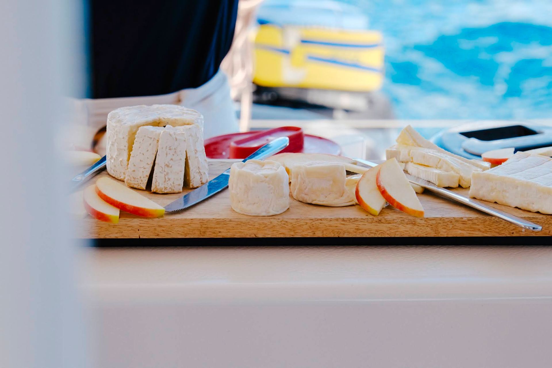 Cheese and apple slices on a wooden board, with a person in the background, near blue water.