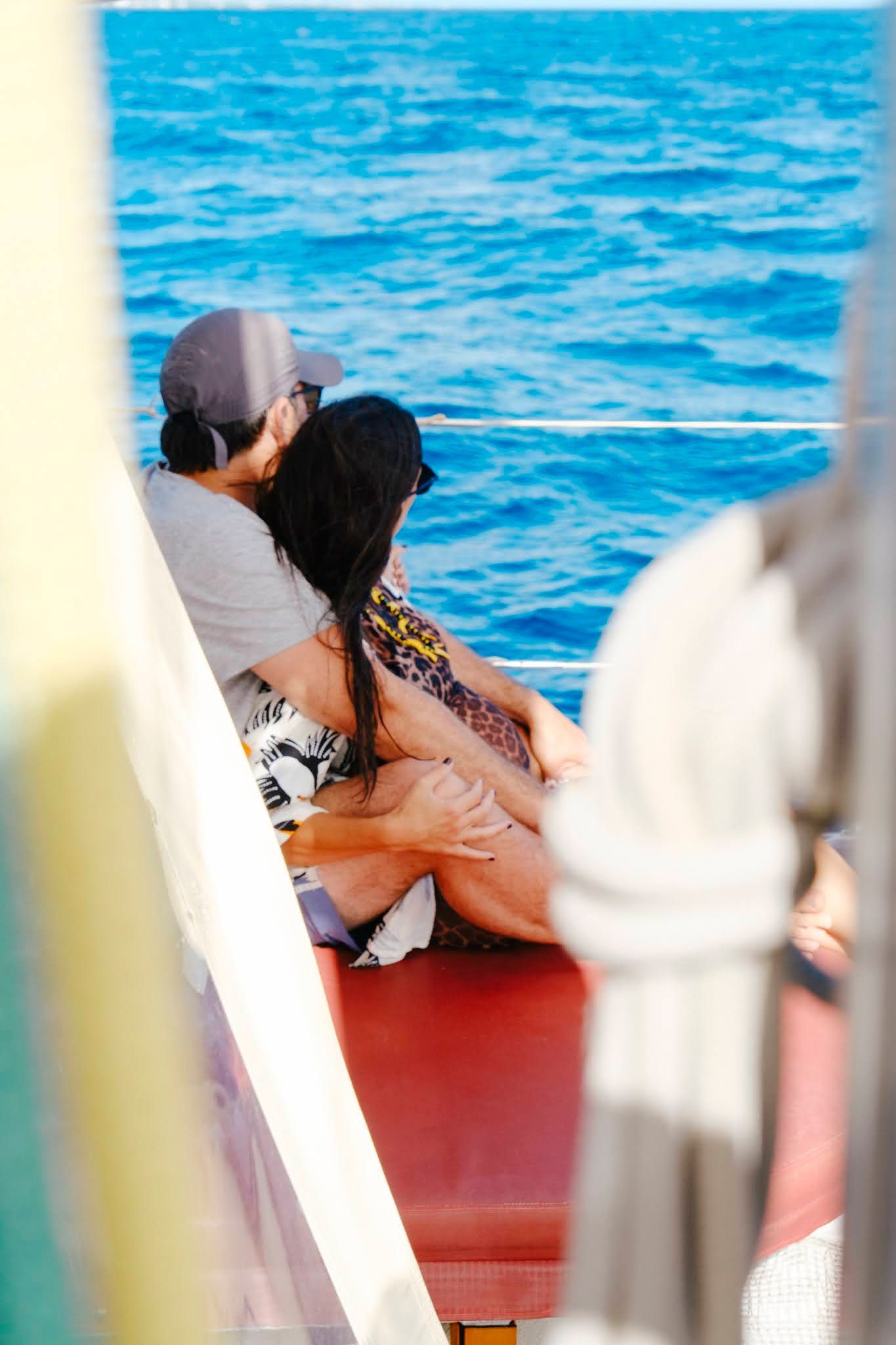 Couple on a boat hugging, facing ocean. Gray cap, floral dress, red bench, blue water.