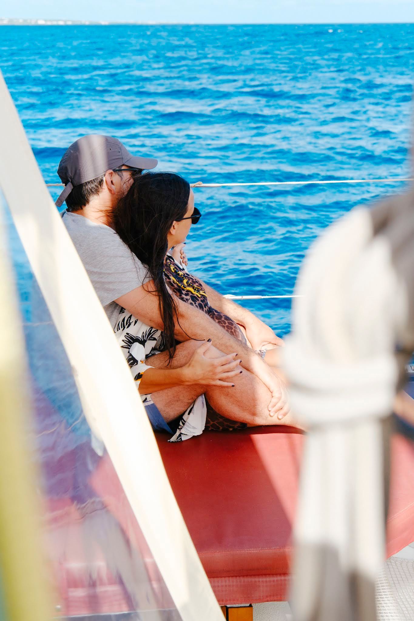 Couple embraces on a boat, gazing at the blue ocean. Red bench visible.