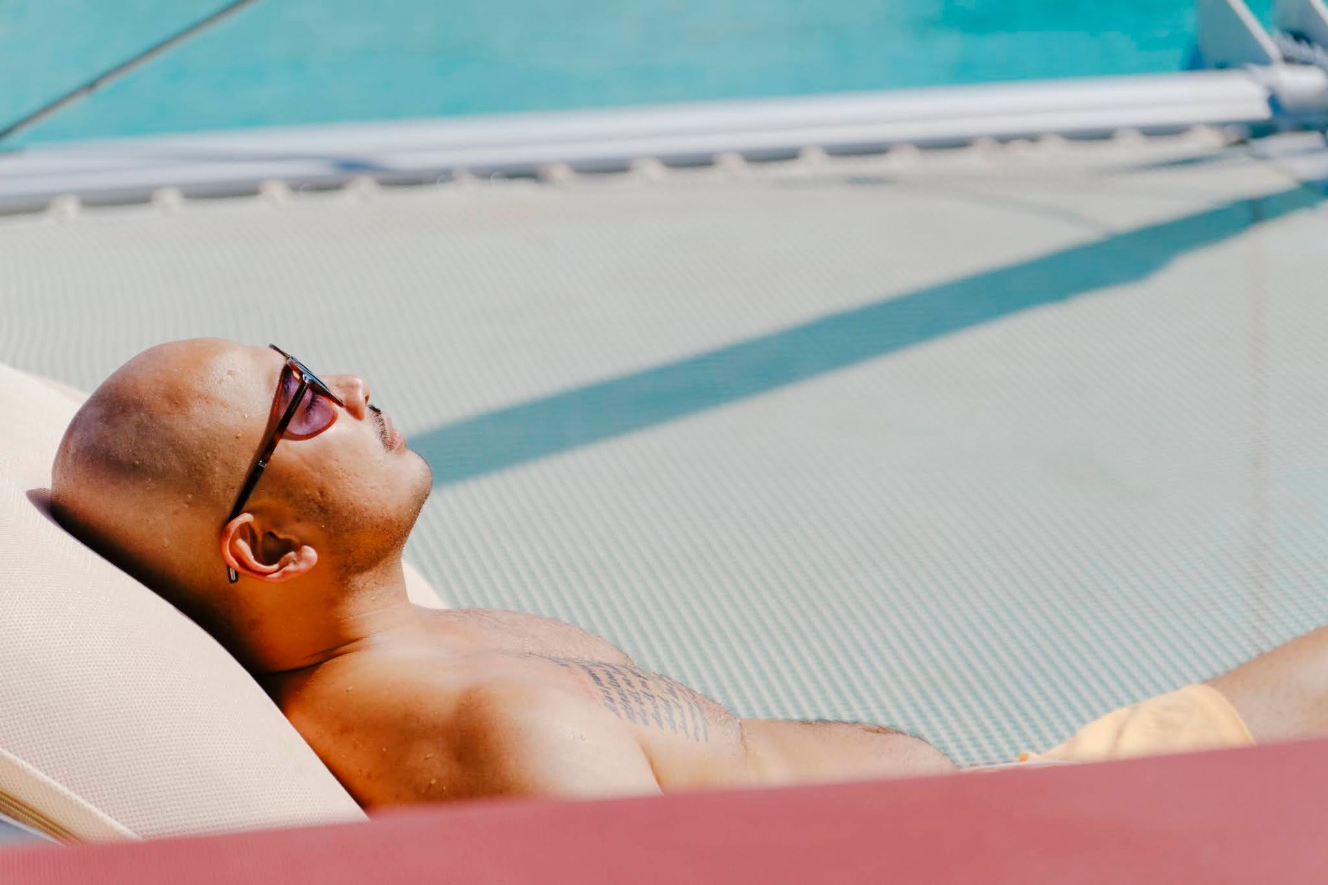 Bald person wearing sunglasses sunbathing on a white mesh surface, near blue water.