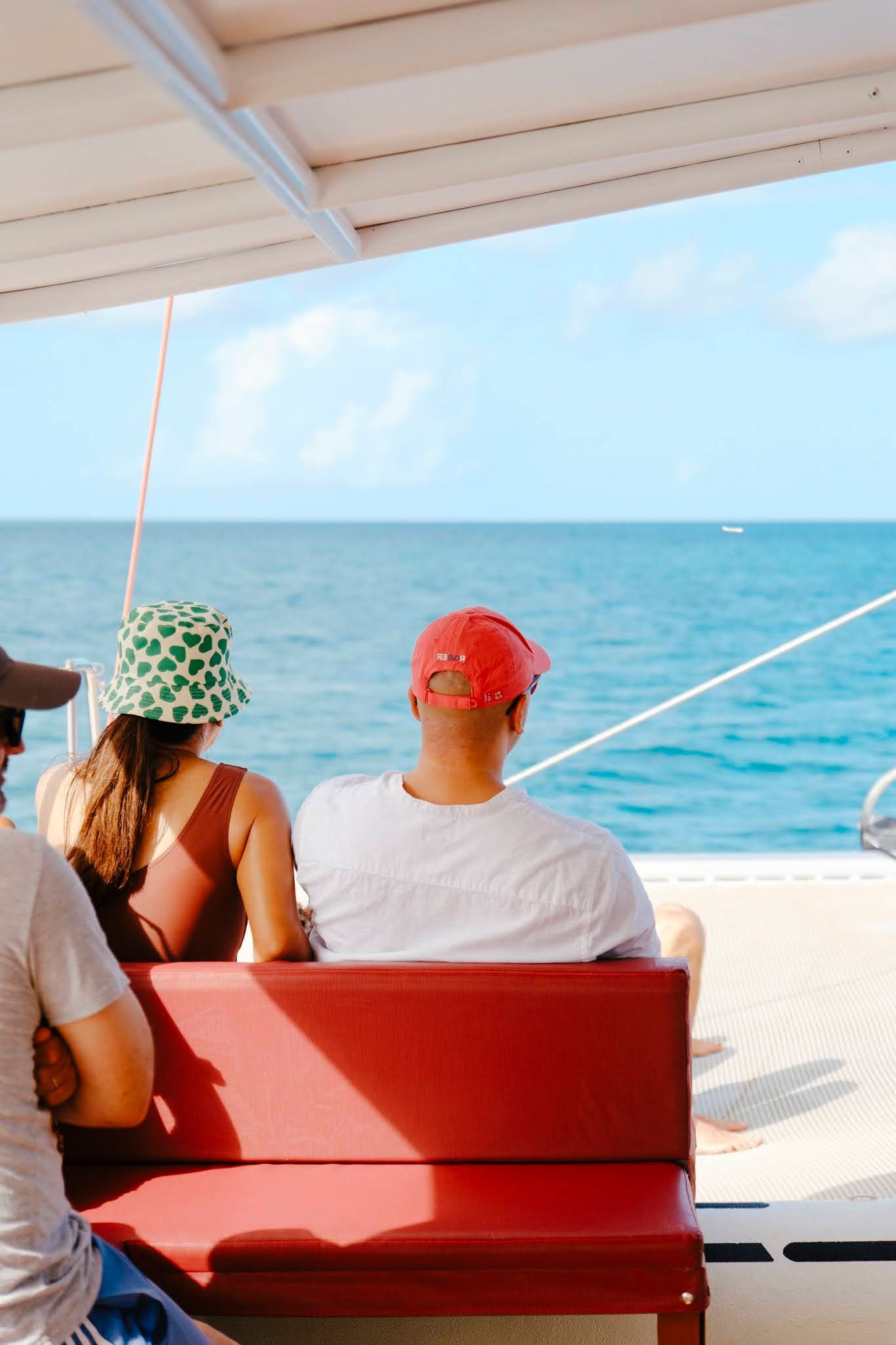 Des personnes sur un bateau contemplent l'océan bleu sous un toit blanc. L'une porte un chapeau rouge, l'autre un chapeau vert.