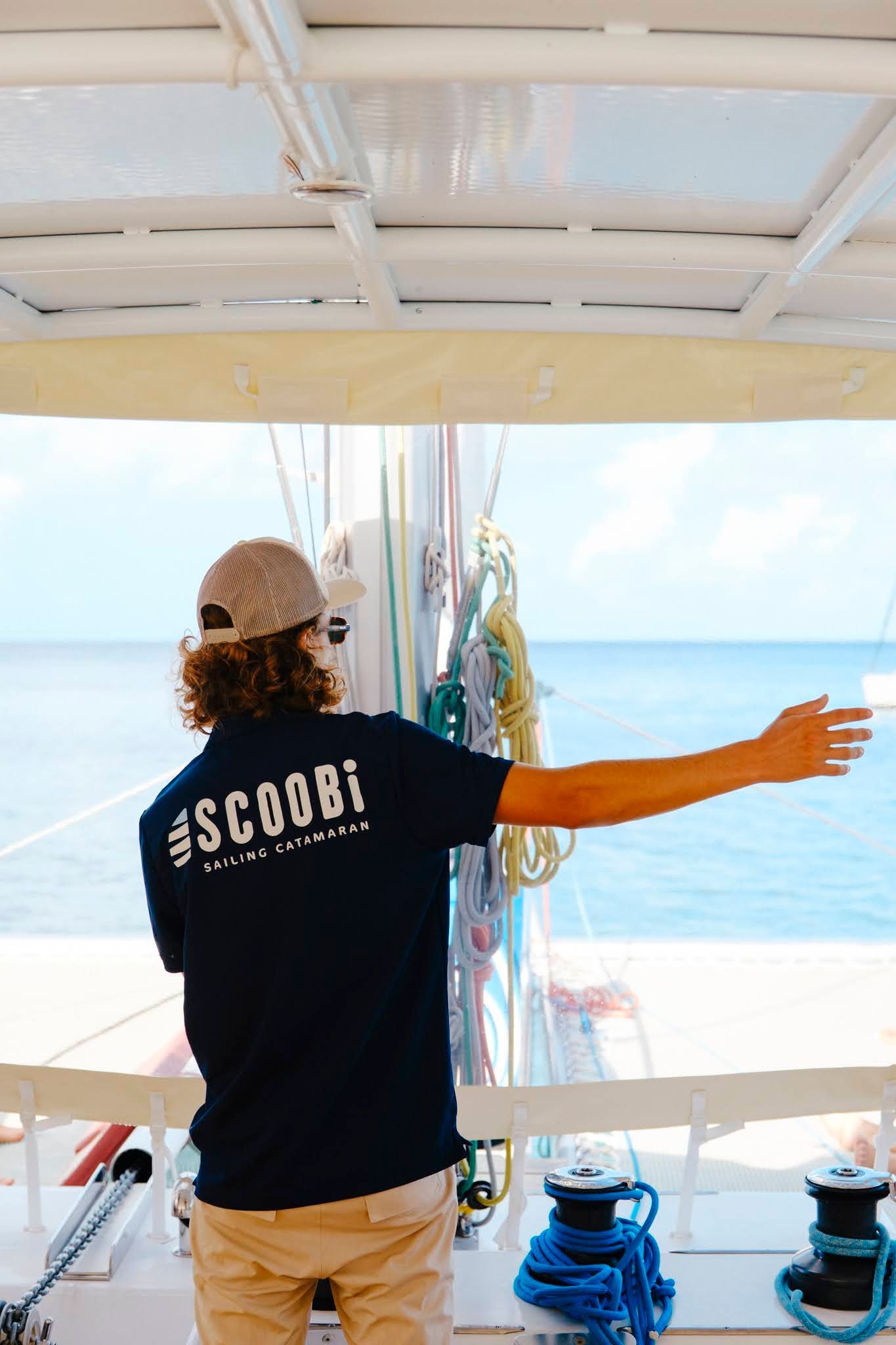 Un homme sur un bateau, pointant du doigt l'océan. Il porte un chapeau et une chemise bleu foncé avec l'inscription « SCOBBI » dans le dos.