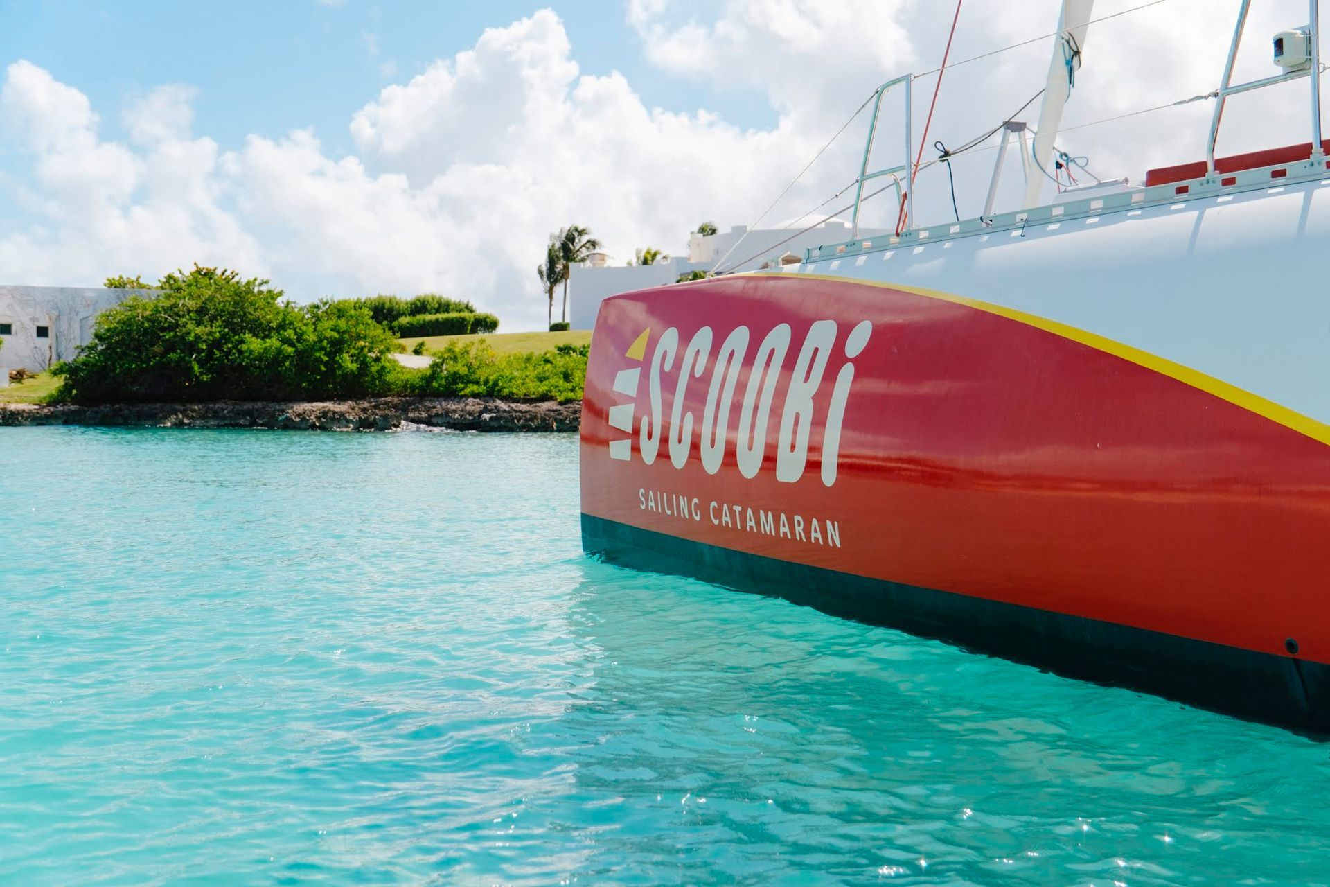 Le catamaran rouge et blanc « Scoobi » sur une eau turquoise, sous un ciel ensoleillé et nuageux.