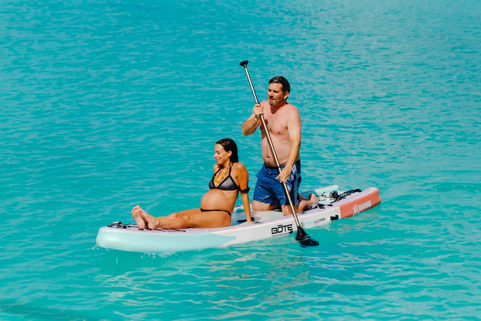 Pregnant woman sits on a paddleboard, man stands and paddles in turquoise water.