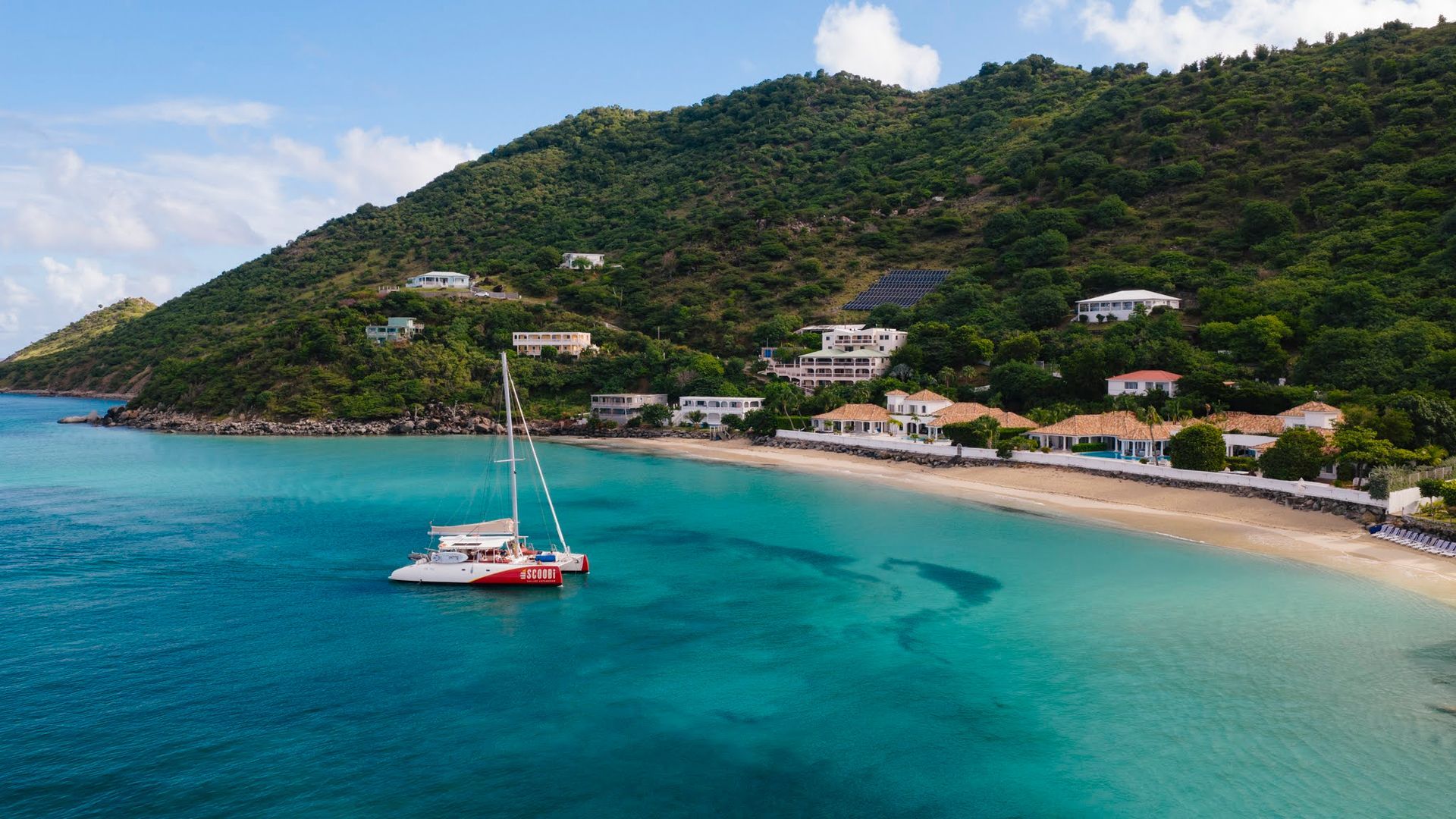 Catamaran in turquoise water near a beach lined with buildings and a lush green hillside under a blue sky.