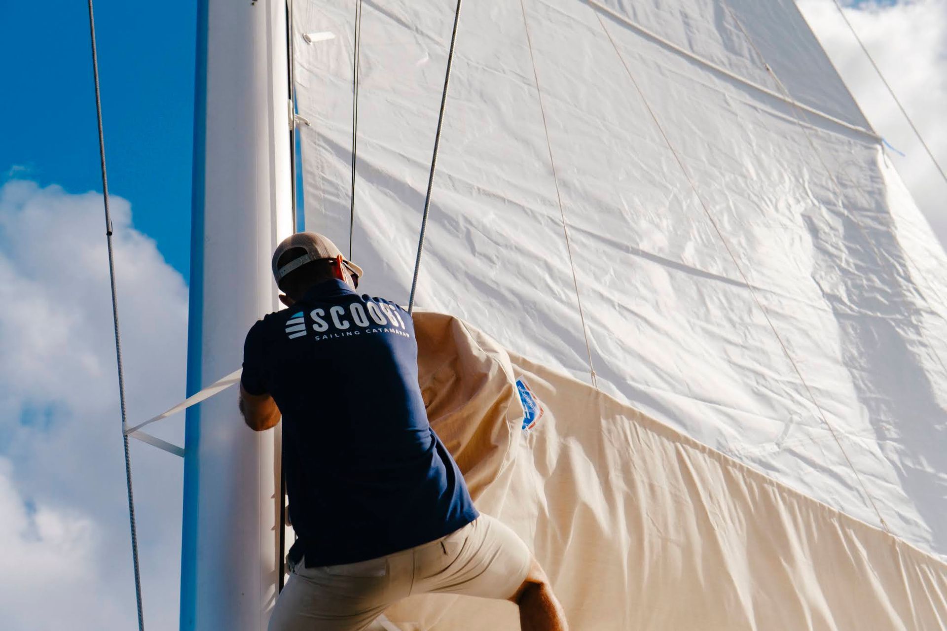 Un homme sur un voilier, fixant la voile au mât sur fond de ciel bleu.