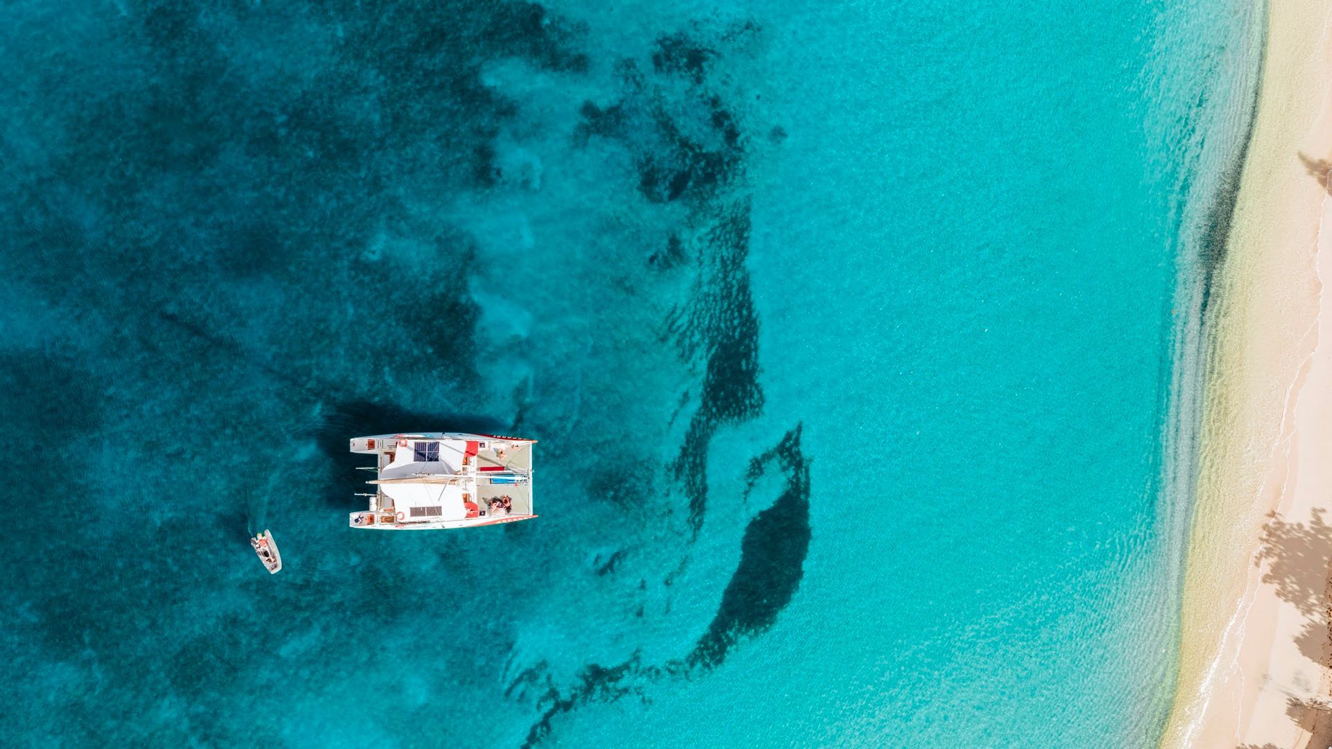 Catamaran et petit bateau dans une eau turquoise près d'une plage de sable.