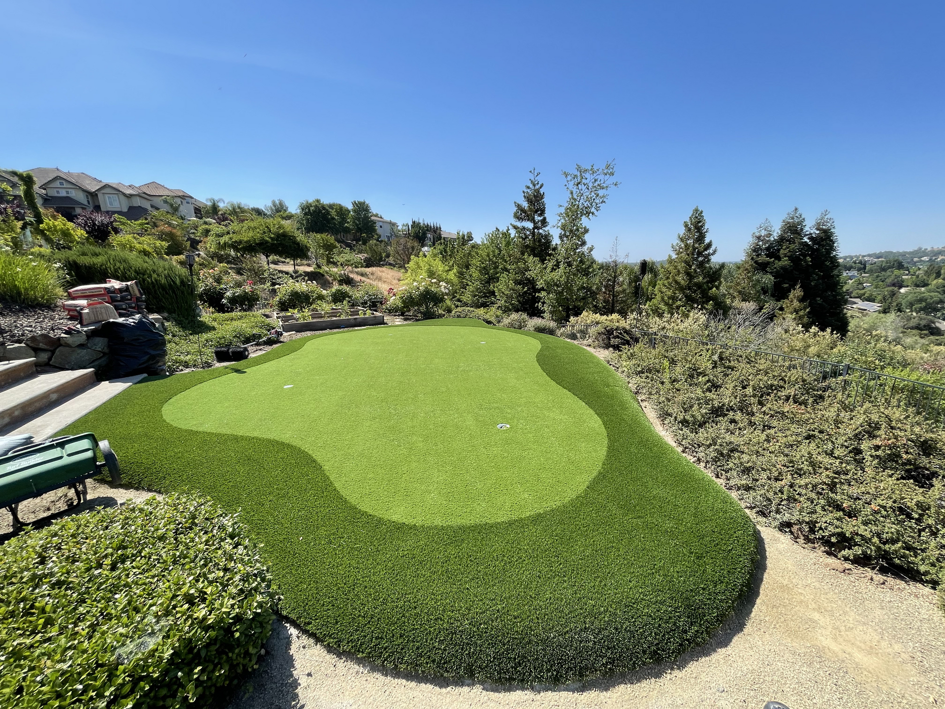 Bright green landscaped mound with a circular lawn pattern in a sunny garden, surrounded by shrubs and gravel paths