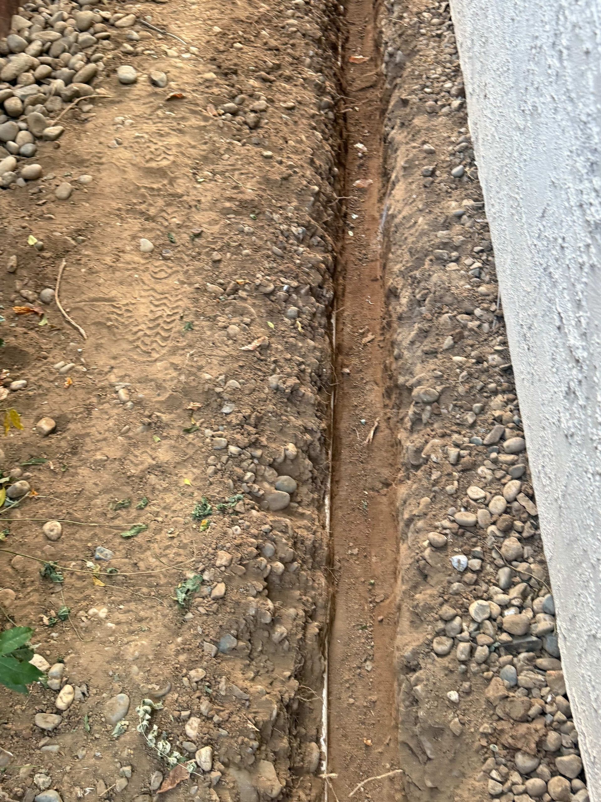 Narrow dirt trench beside a concrete wall, with loose soil and small rocks along the edge