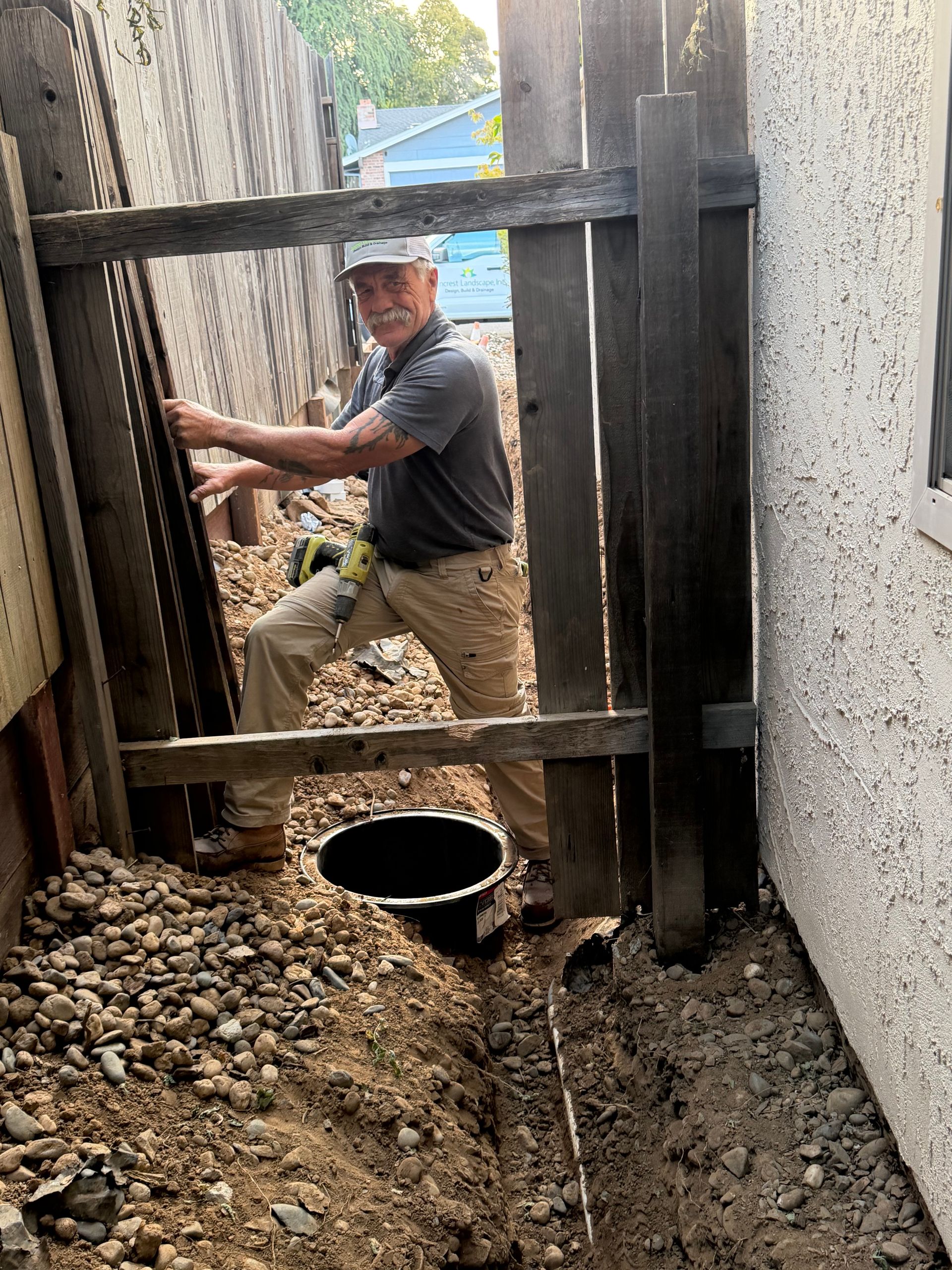 Worker on muddy trench floor installing a black pipe beside a house foundation.