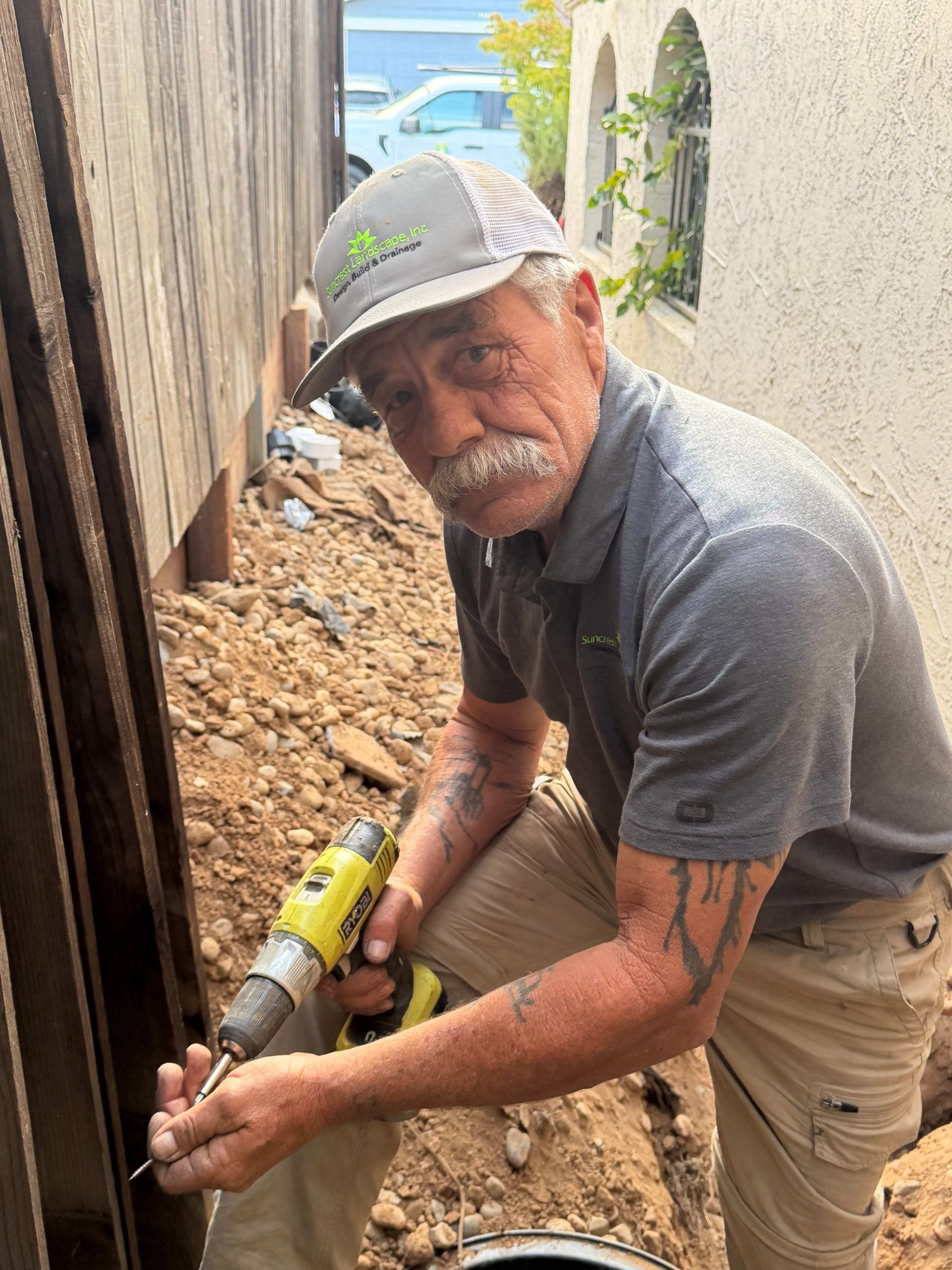 Worker in gray shirt and cap drilling beside a wooden fence in a narrow dirt trench.