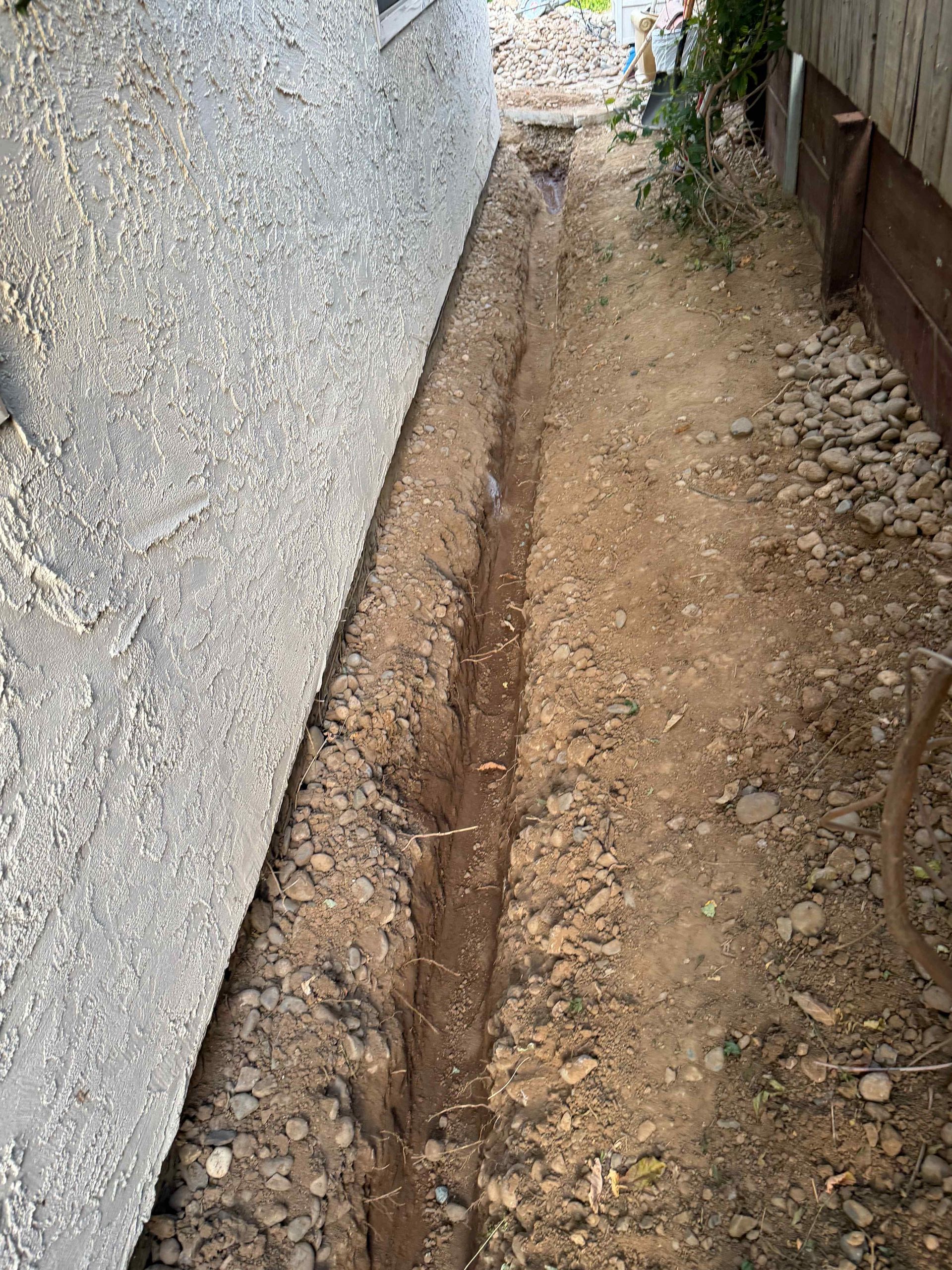 Narrow dirt trench along a wall with exposed pipe or cable running through it.