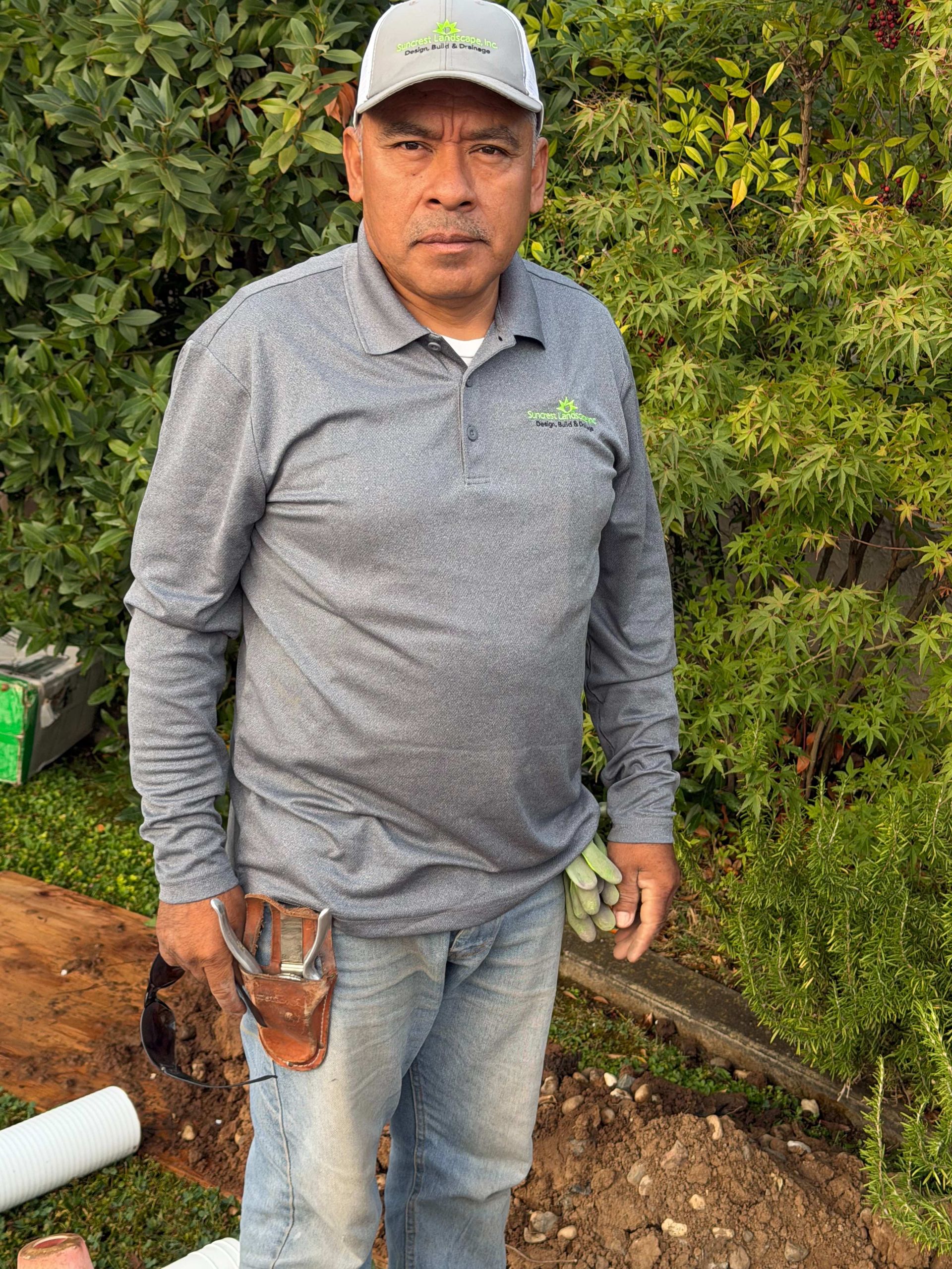 Man standing outdoors in a gray shirt and cap, holding a tool in a garden area.