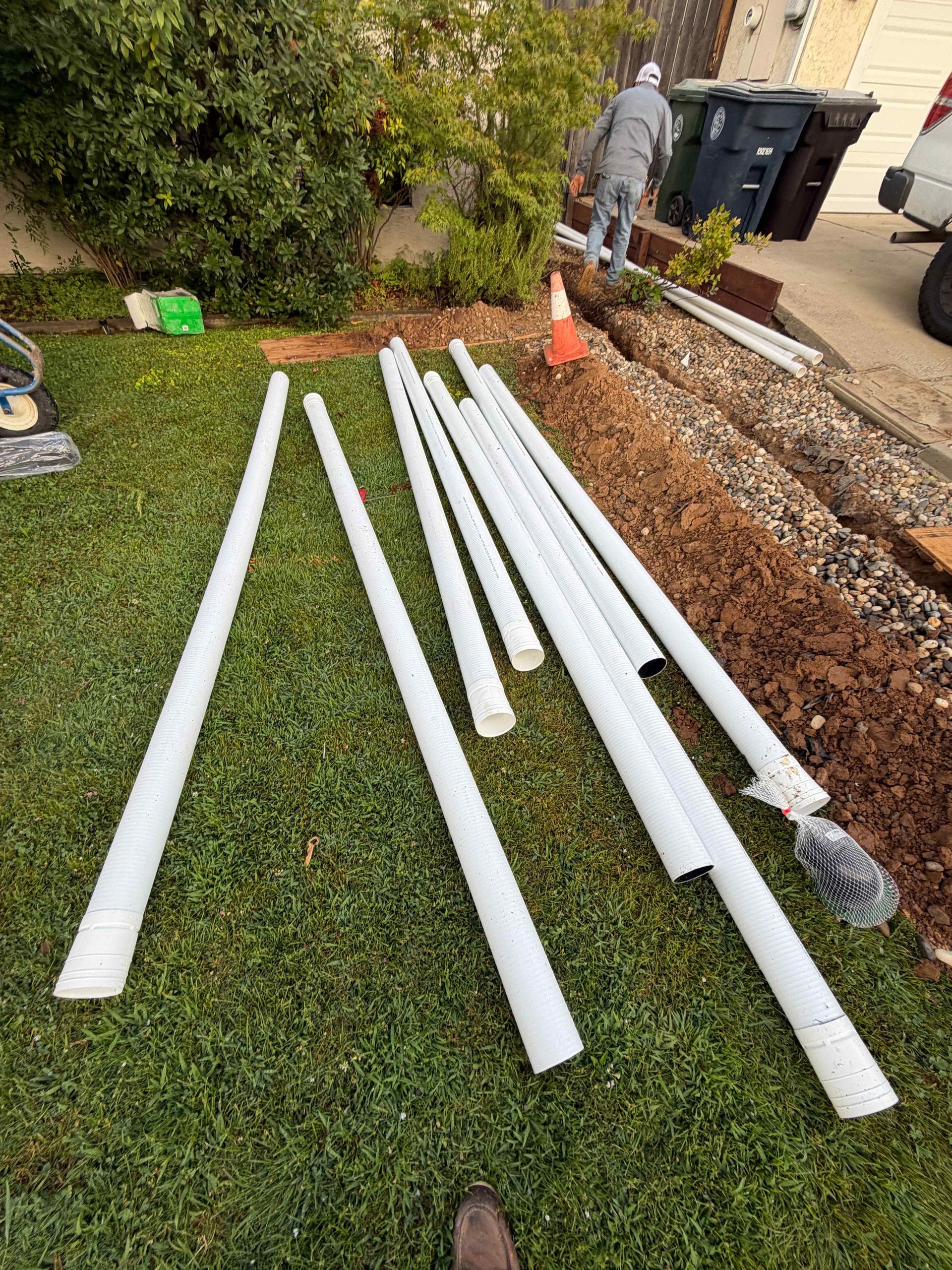 White PVC pipes laid on grass in a yard next to a walkway and trash bins