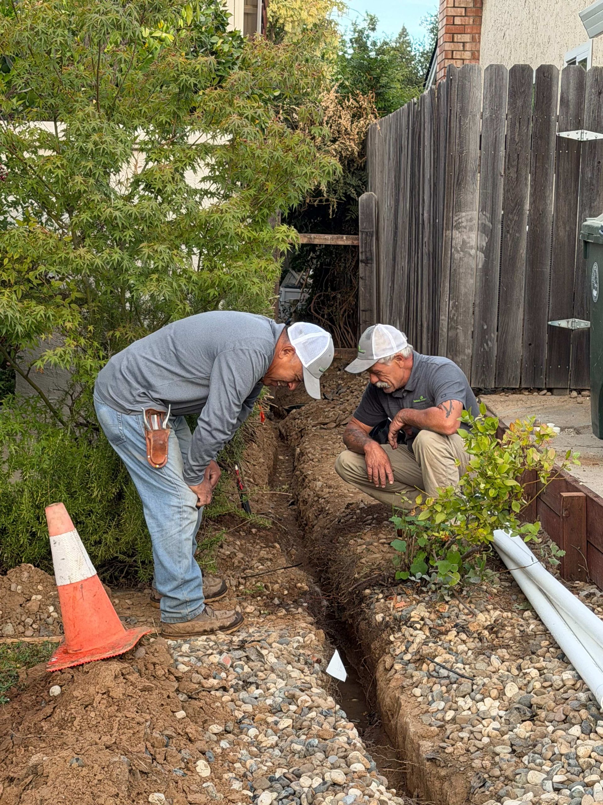 Two workers in white caps dig a trench beside a wooden fence and house, with an orange cone nearby.
