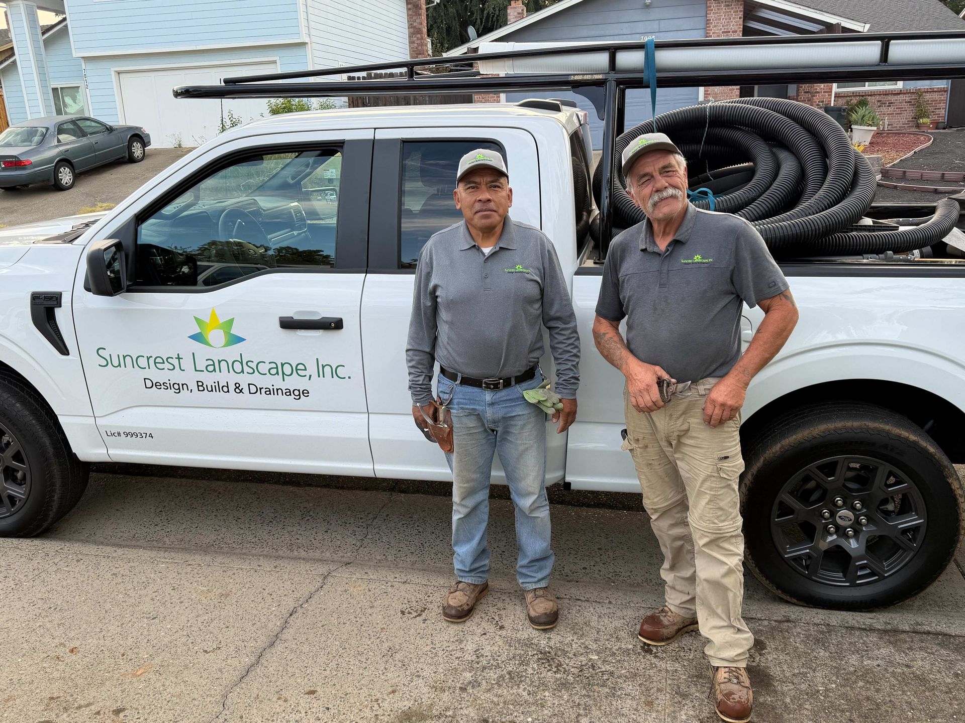 Two workers stand beside a white service truck with tools and coiled hose in the back.