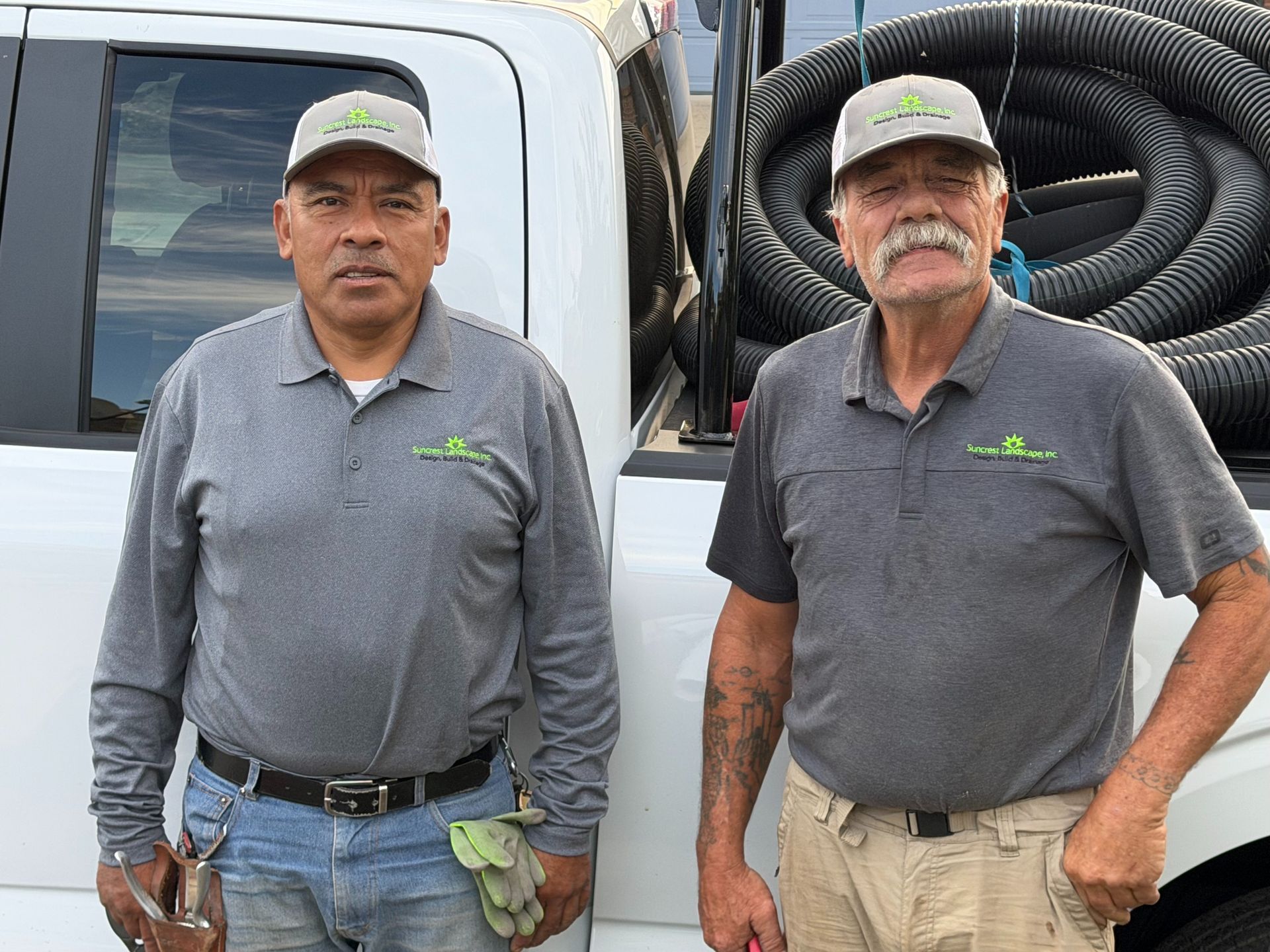 Two men in gray shirts stand by a truck with coiled hoses in the back.