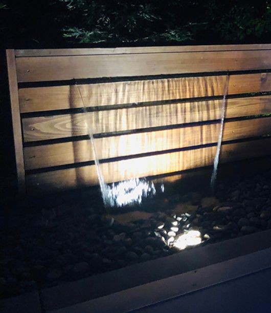 Nighttime wooden fence lit by a spotlight, with a small ground light on rocks below.
