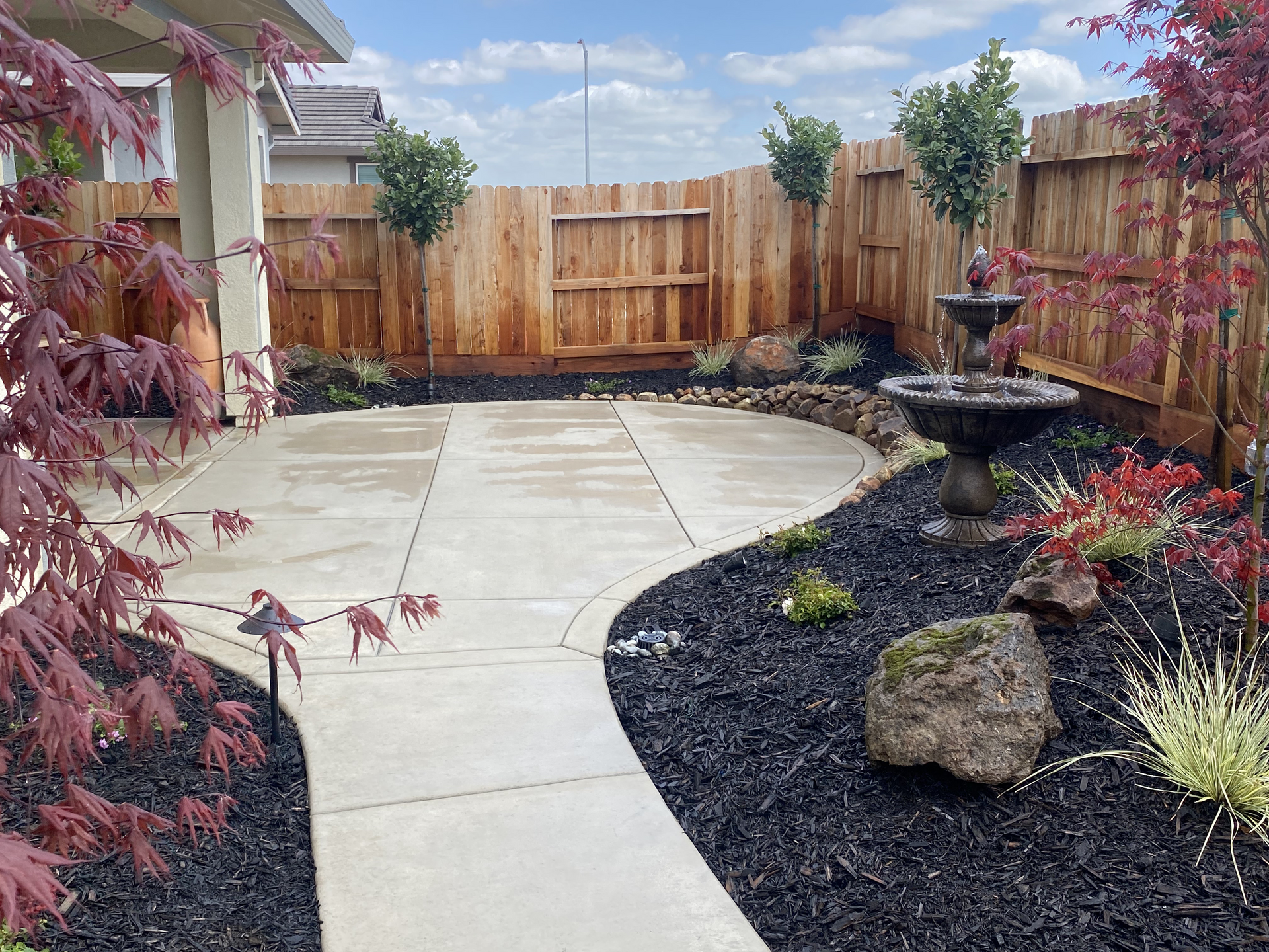 Curved concrete patio beside black gravel garden, wooden fence, fountain, and colorful shrubs in a backyard