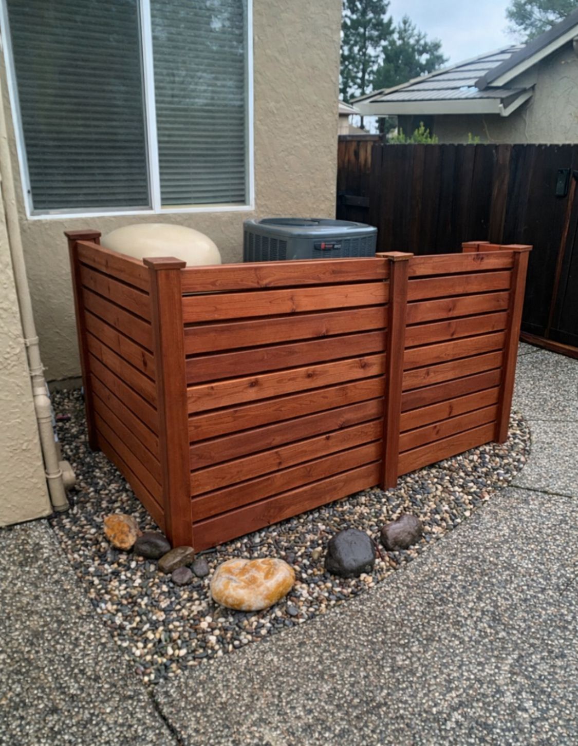 Wooden privacy screen around outdoor HVAC unit beside a house and gravel patio area