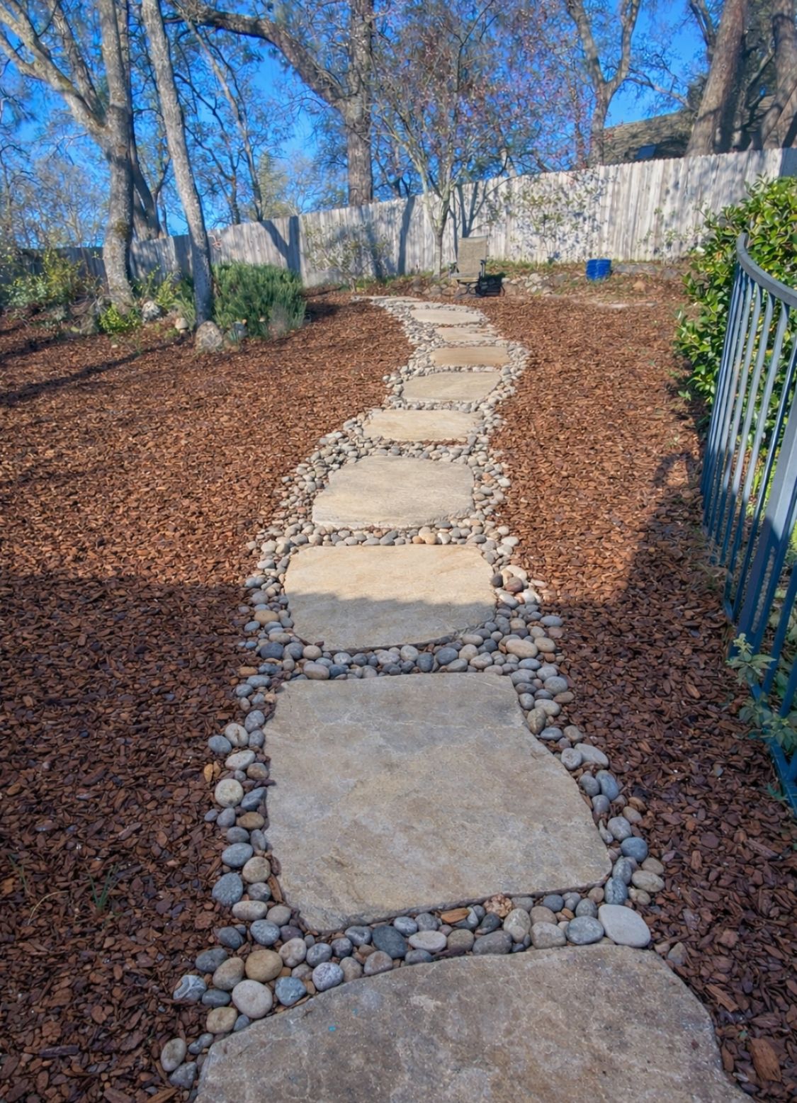 Stepping-stone path through a mulched garden with bare trees and a stone wall on a sunny day