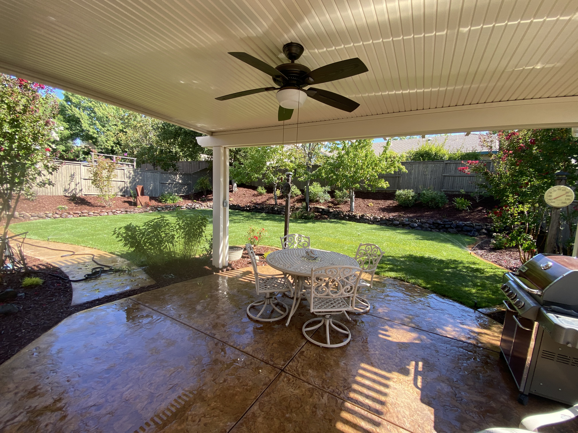 Covered backyard patio with ceiling fan, table, grill, and wet concrete overlooking a green garden