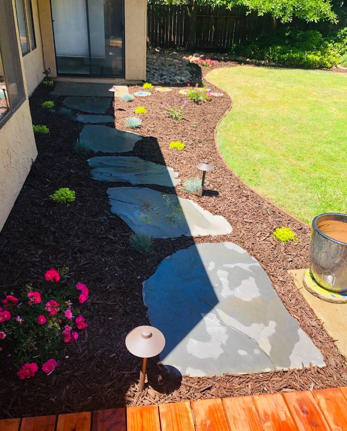 Stone path through a landscaped front yard with red flowers, mulch, green lawn, and a porch light.