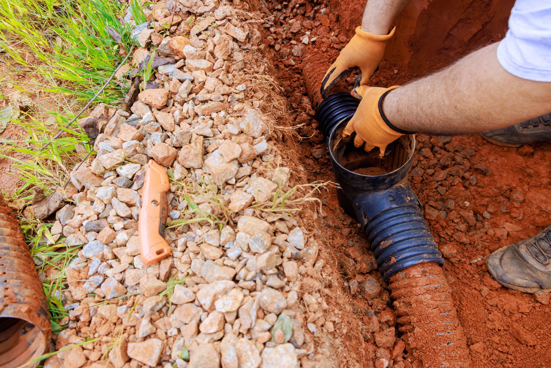 Worker fitting a black drainage pipe into a trench with gravel and orange piping on both sides