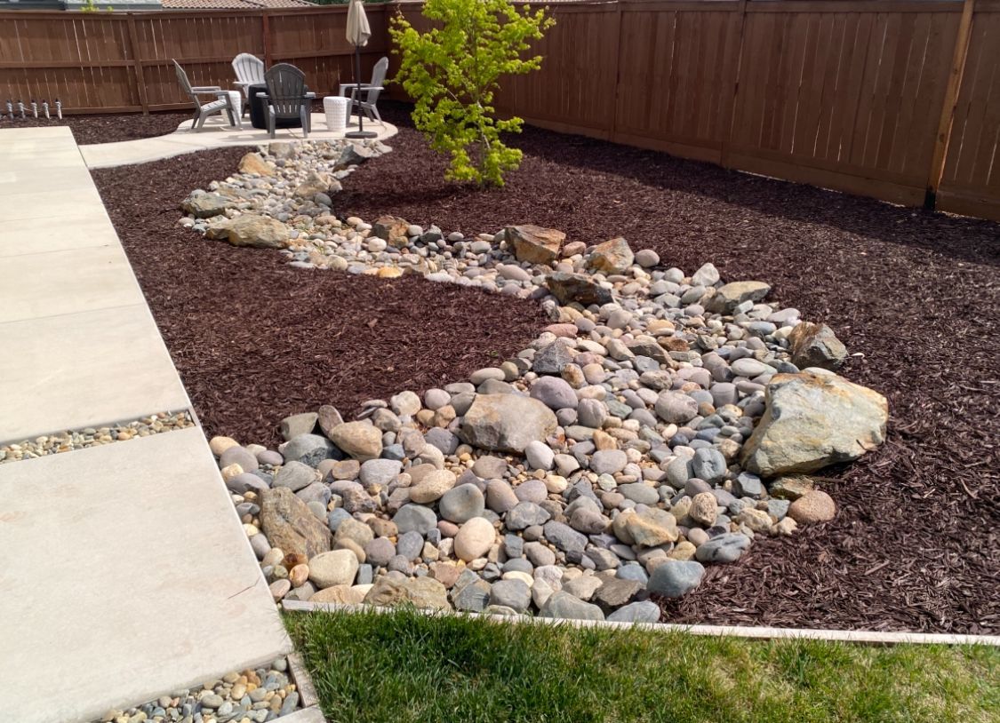 A rock-lined landscape bed beside a concrete walkway and wooden fence, with brown mulch and a green lawn edge.