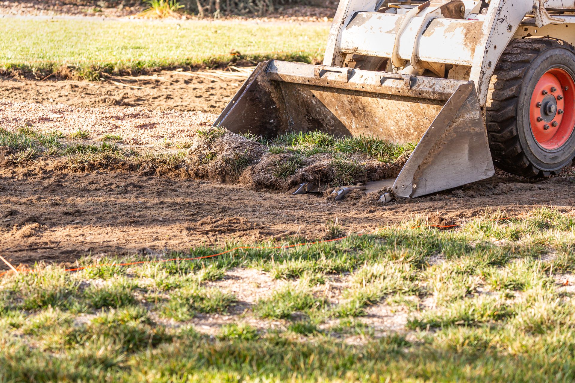 Front loader bucket clearing dirt on a muddy yard beside grass