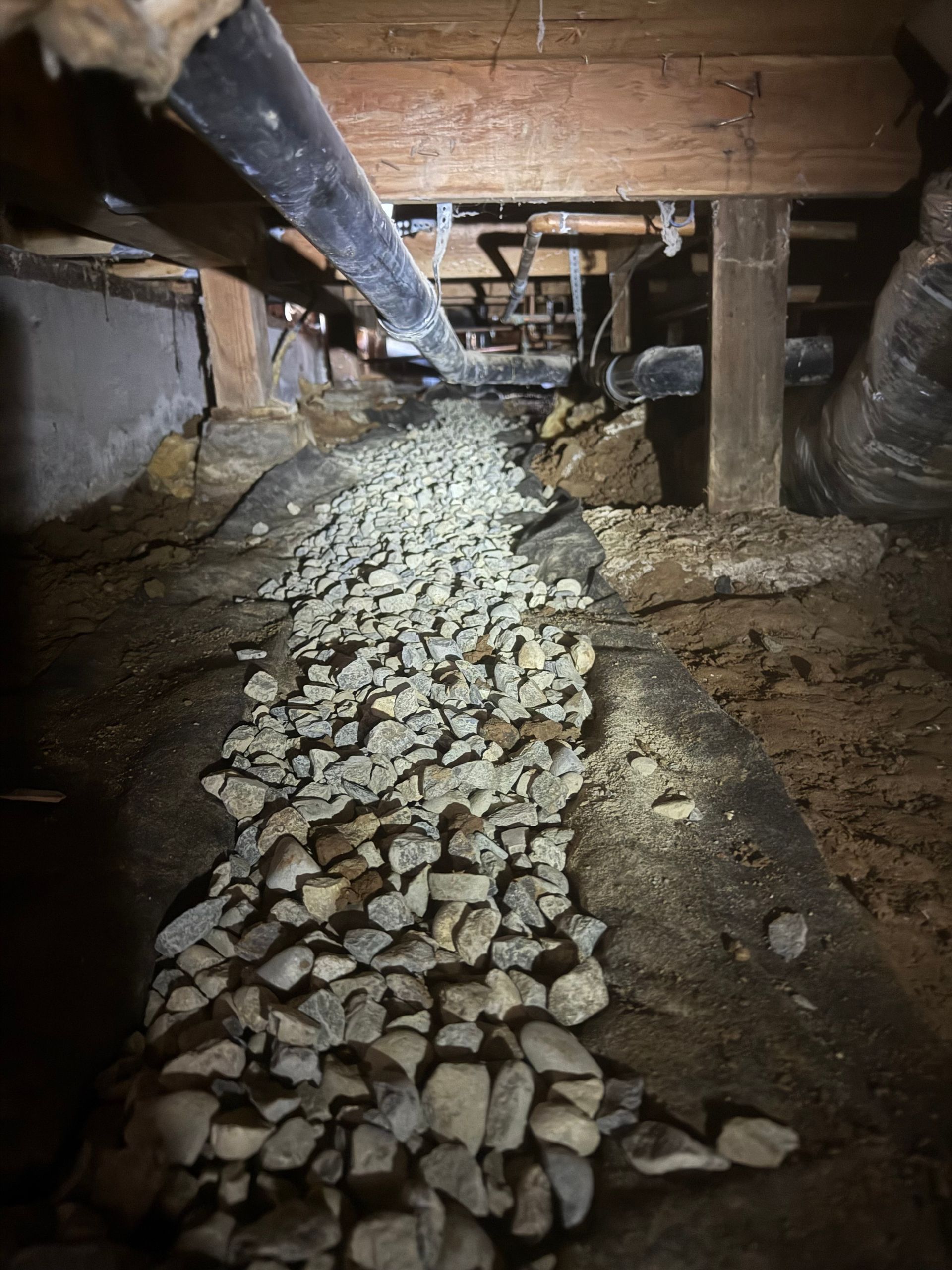 Rock-lined crawlspace under a wooden floor with exposed beams and pipes overhead