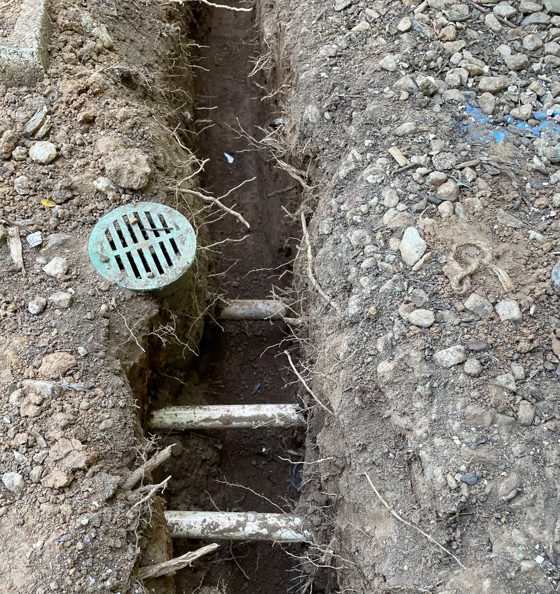 Narrow trench with exposed metal rungs and a small green drain grate in dirt and gravel