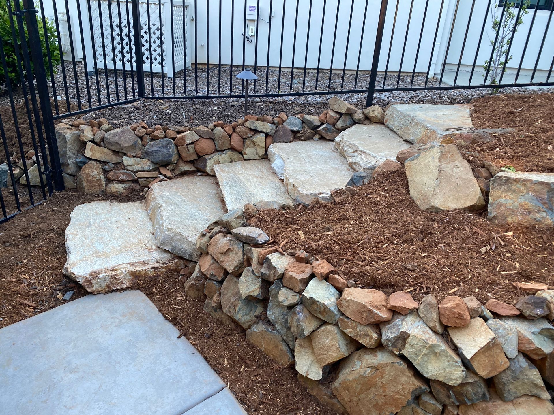 Stone steps and retaining wall in a rocky yard beside a black metal fence