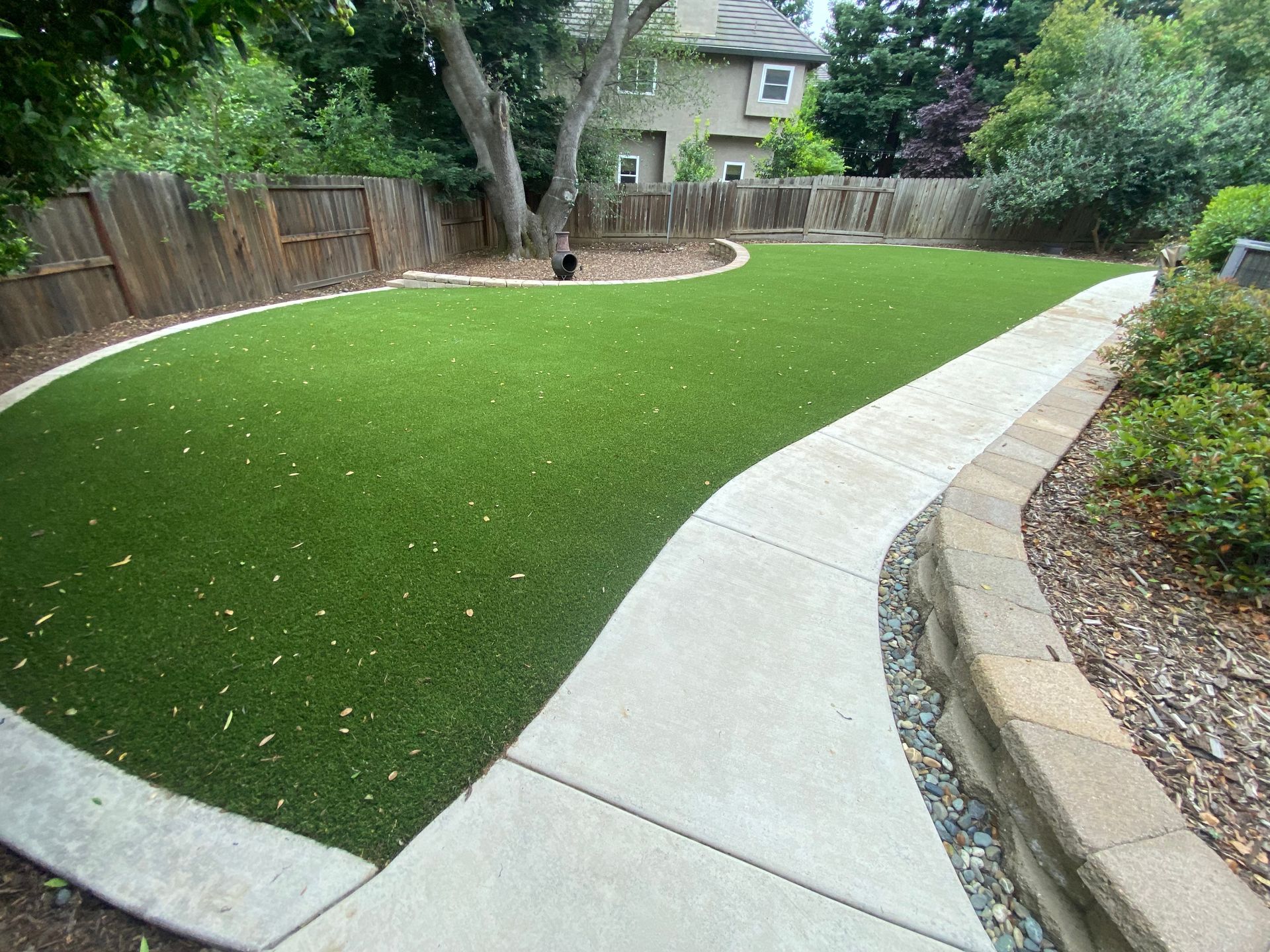 Backyard with curved concrete walkway around a green lawn, wooden fence, and trees