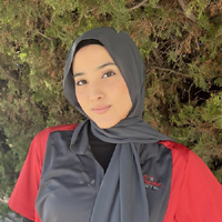 A woman wearing a hijab and a red shirt is standing in front of a tree.