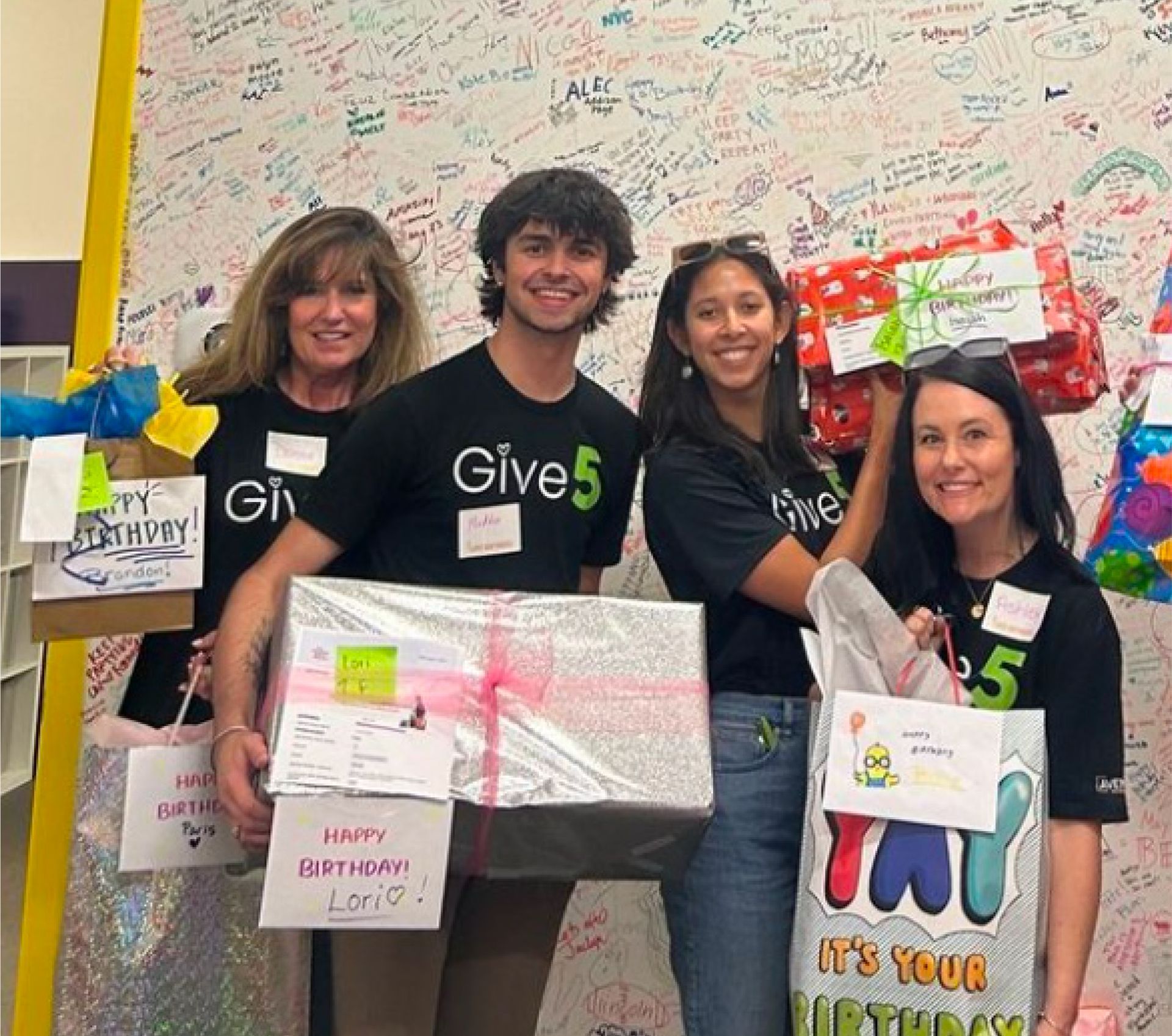 Four volunteers in black “Give” shirts hold wrapped gifts in front of a message-covered wall.